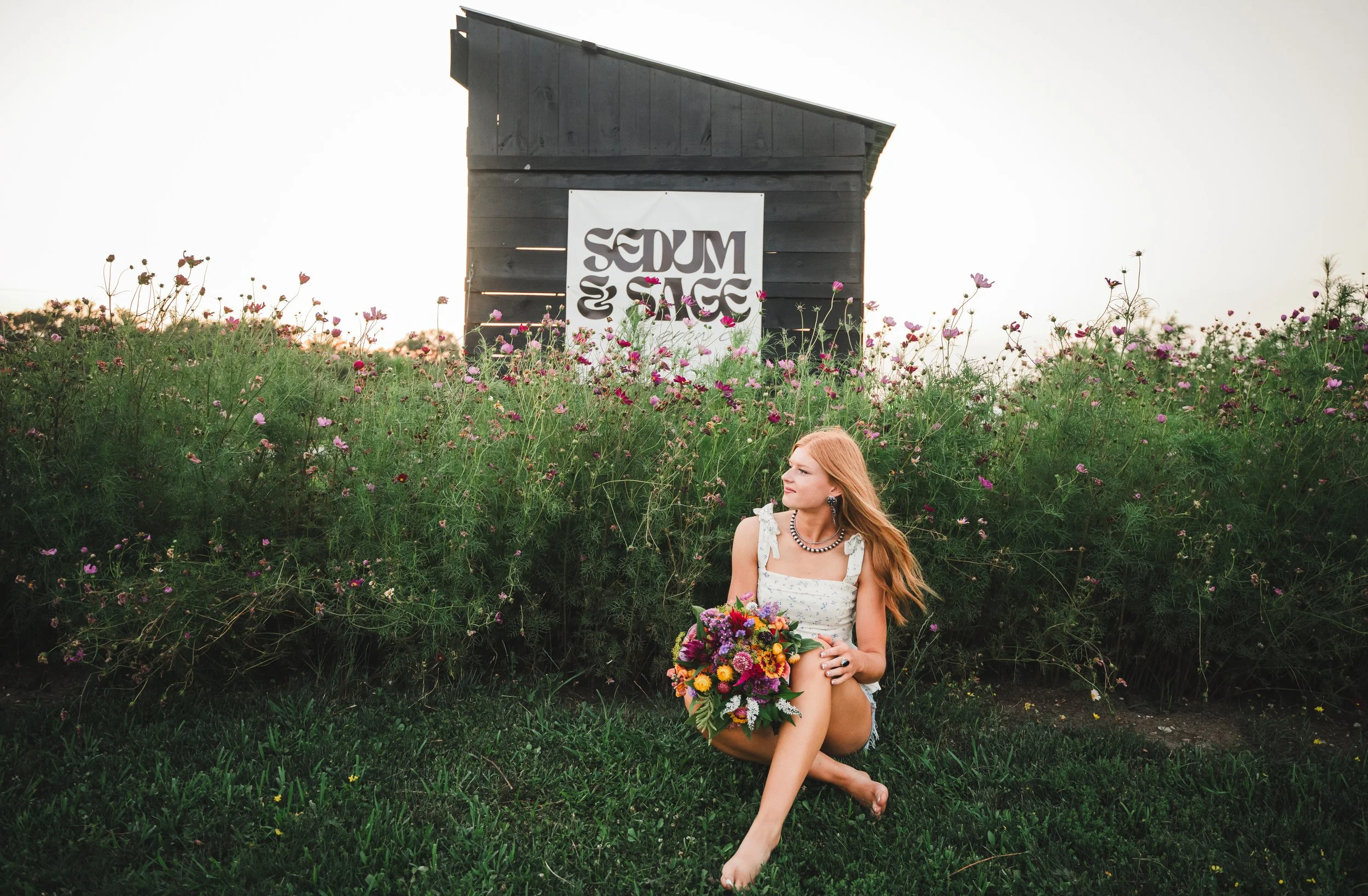 A woman with long blonde hair sitting on grass in front of a bush with pink and purple flowers, holding a colorful bouquet of flowers, with a black wooden structure and a white sign with black writing in the background.