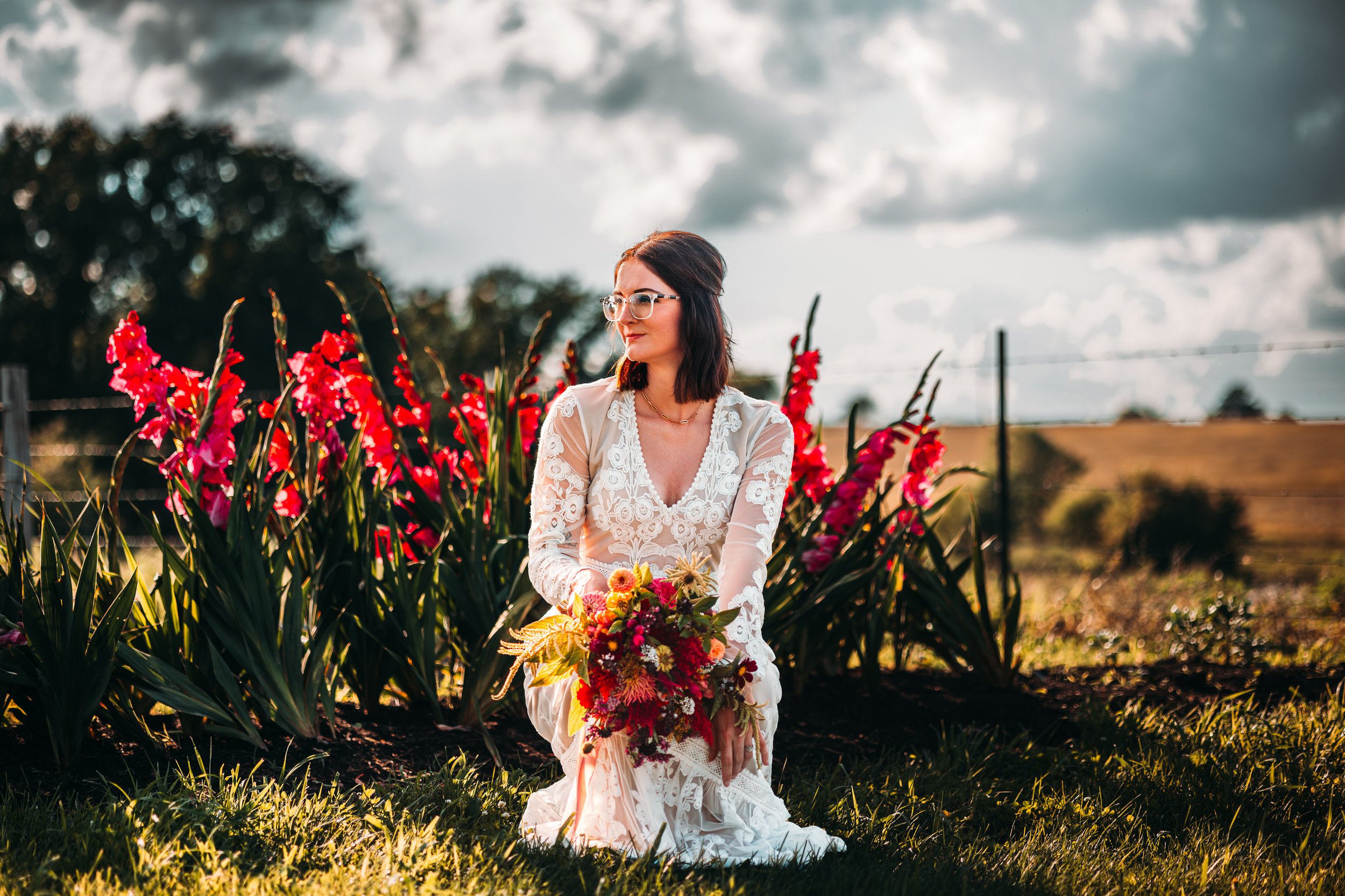A woman in a white lace dress sitting on grass, holding a bouquet of flowers, surrounded by tall pink flowers and greenery, under a cloudy sky during sunset.