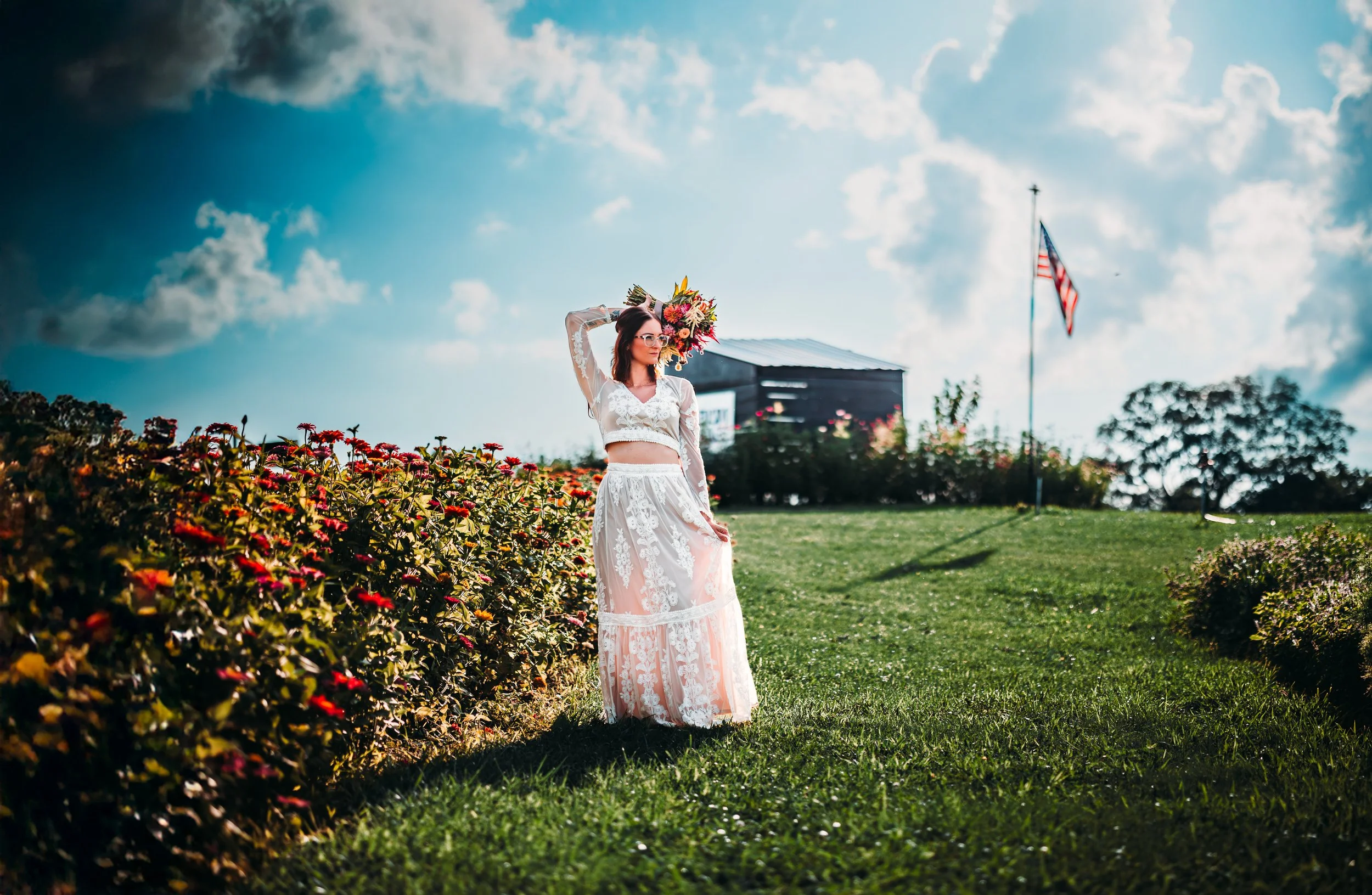A woman in a white lace two-piece dress holds a large colorful bouquet of flowers on a grassy field with flowers and an American flag in the background, under a partly cloudy sky.
