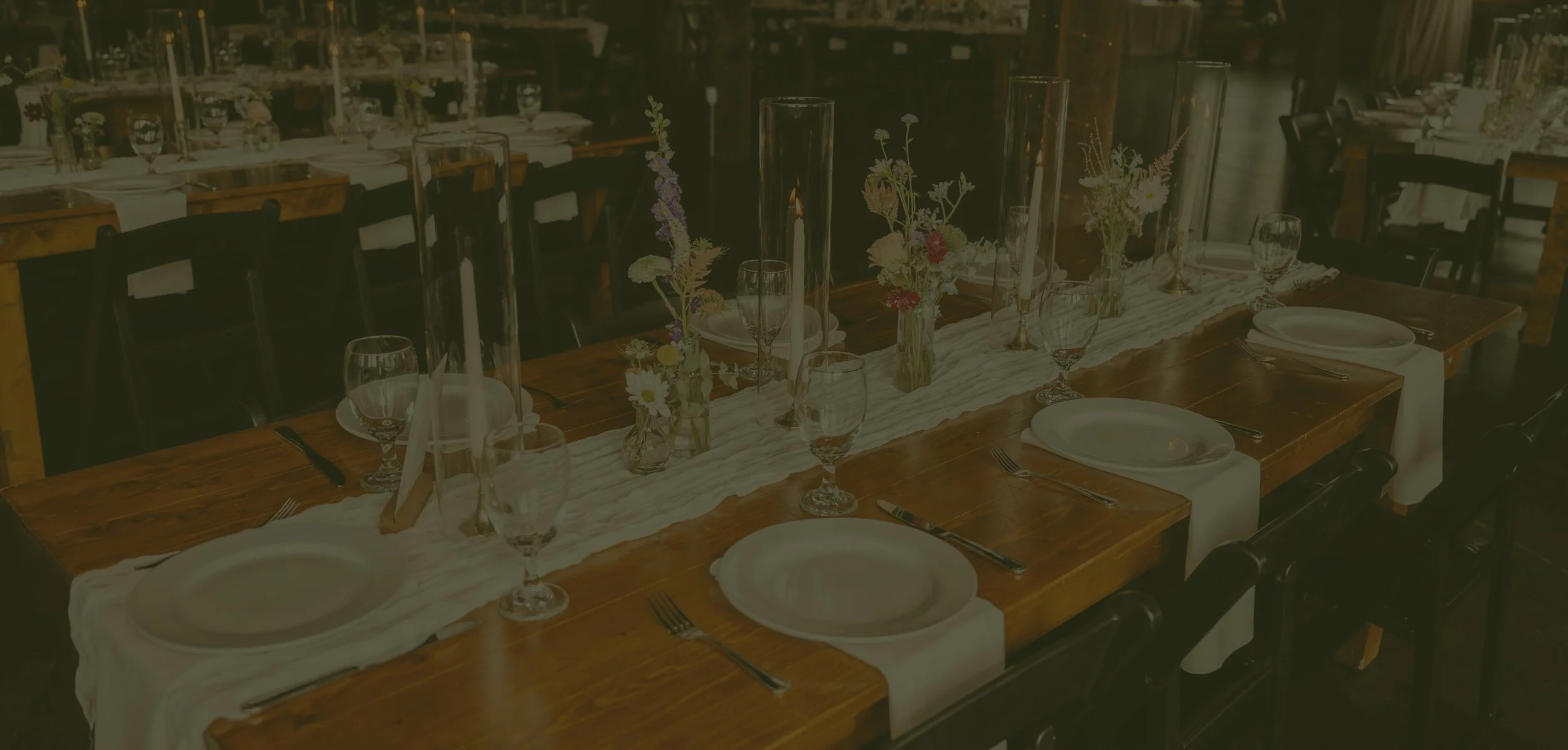 A long wooden banquet table set for a special occasion with white plates, silverware, glasses, and white napkins. The table has a white table runner and centerpieces with small vases of wildflowers and tall candles in glass holders. The room appears to be a rustic dining space with other tables in the background.