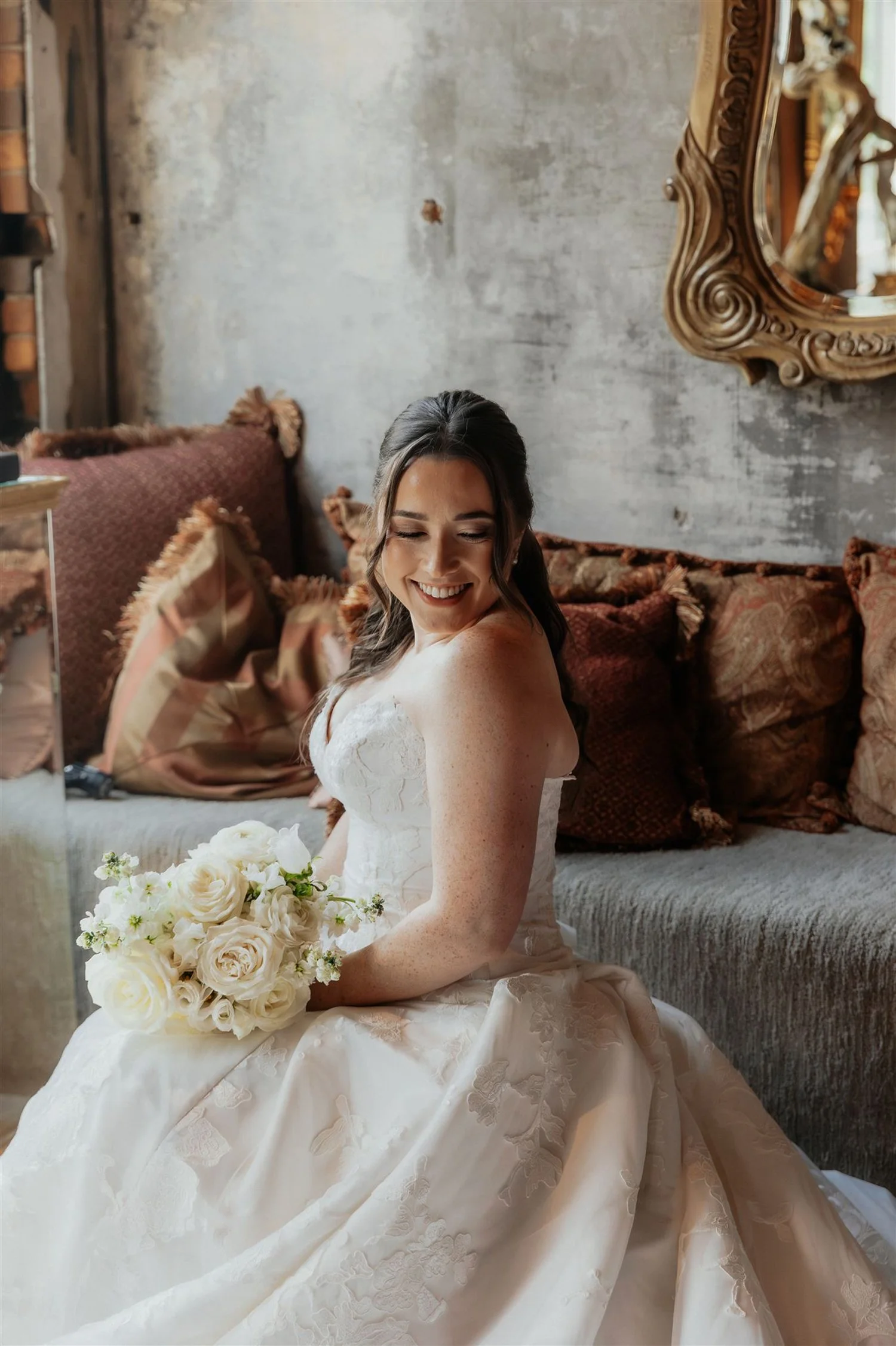 A smiling bride in a white wedding dress holding a bouquet of white roses, sitting in a cozy room with a textured wall and vintage-style cushions.