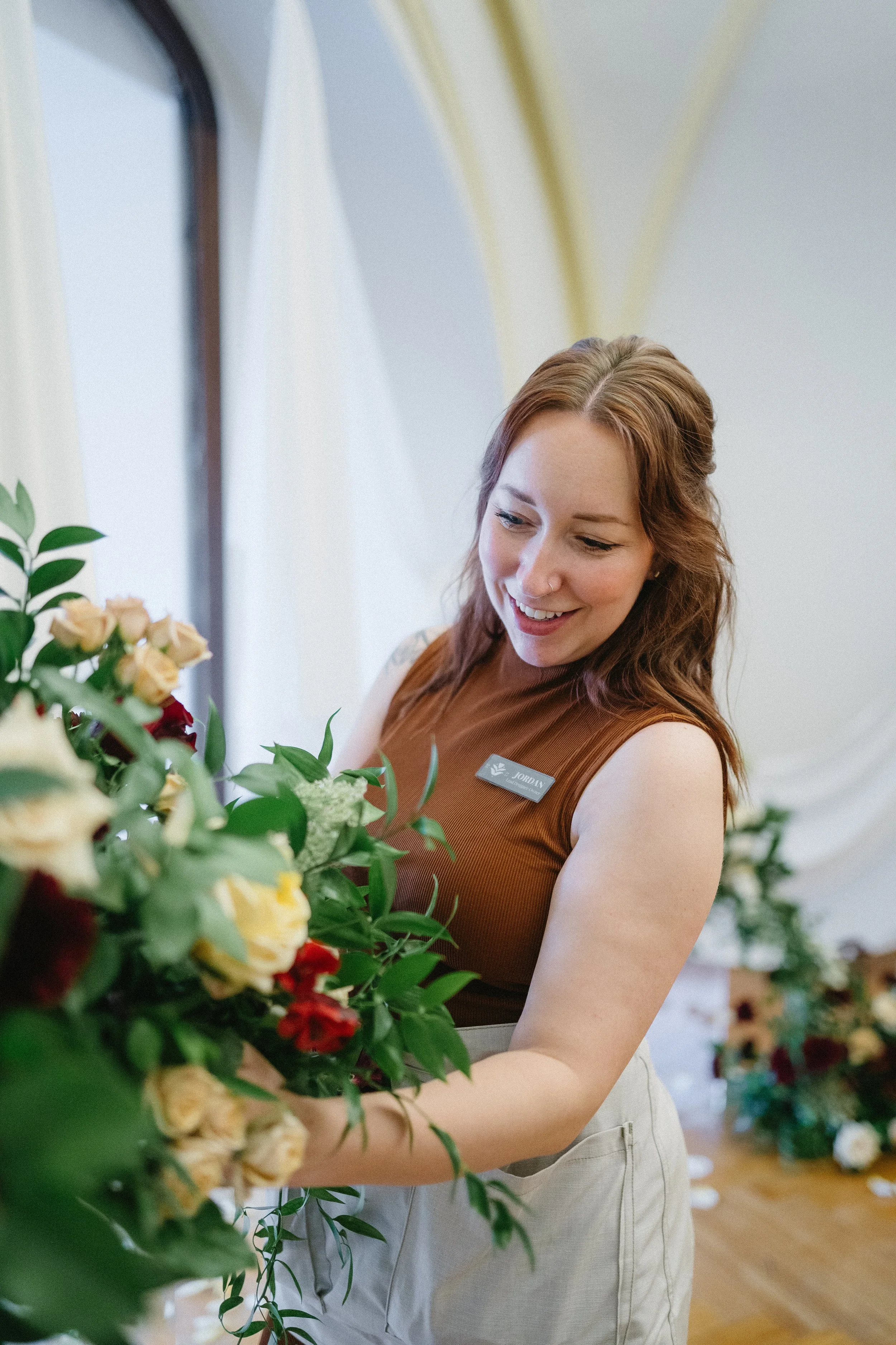 Jordan Schuerman of For Honey Florals arranging flowers indoors, wearing a brown sleeveless top with a name tag, smiling as she works with a bouquet.
