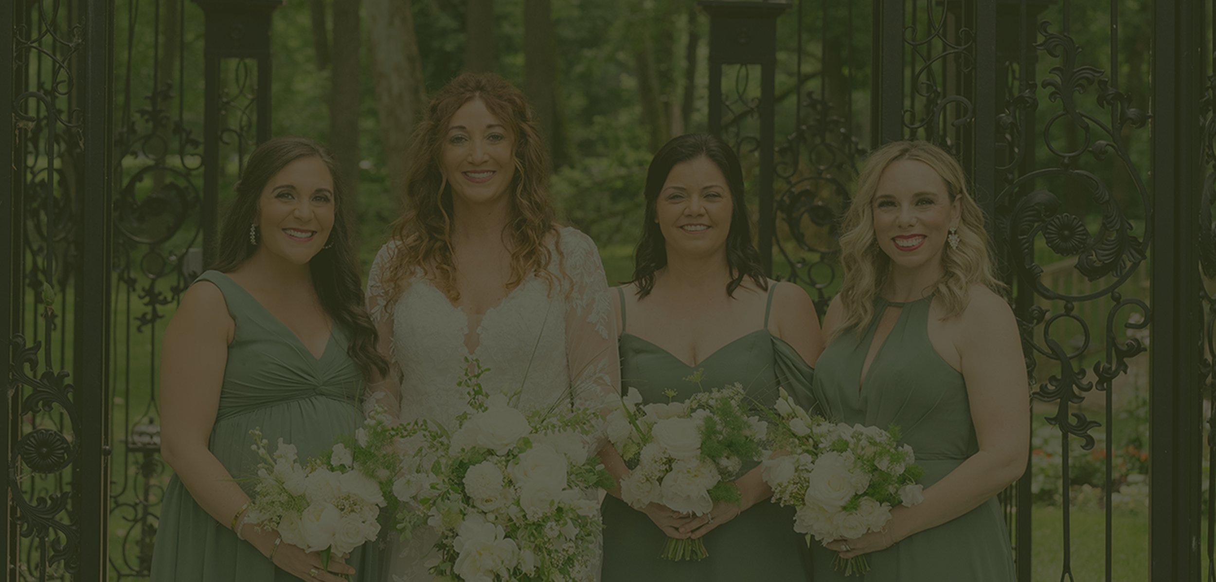 Group of five women dressed in green and white, standing outdoors in front of an ornate metal gate, at what appears to be a wedding or formal event.