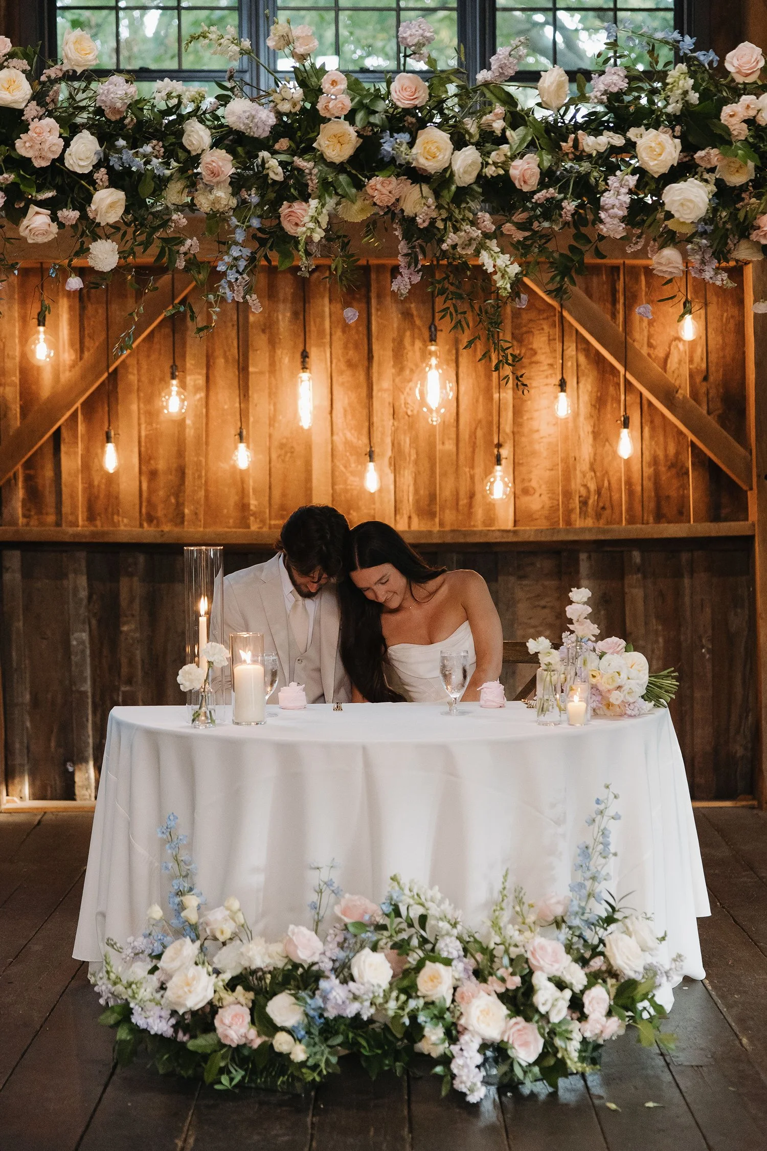 A bride and groom at their wedding table, surrounded by floral arrangements and candles, with a wooden backdrop and hanging Edison bulbs.