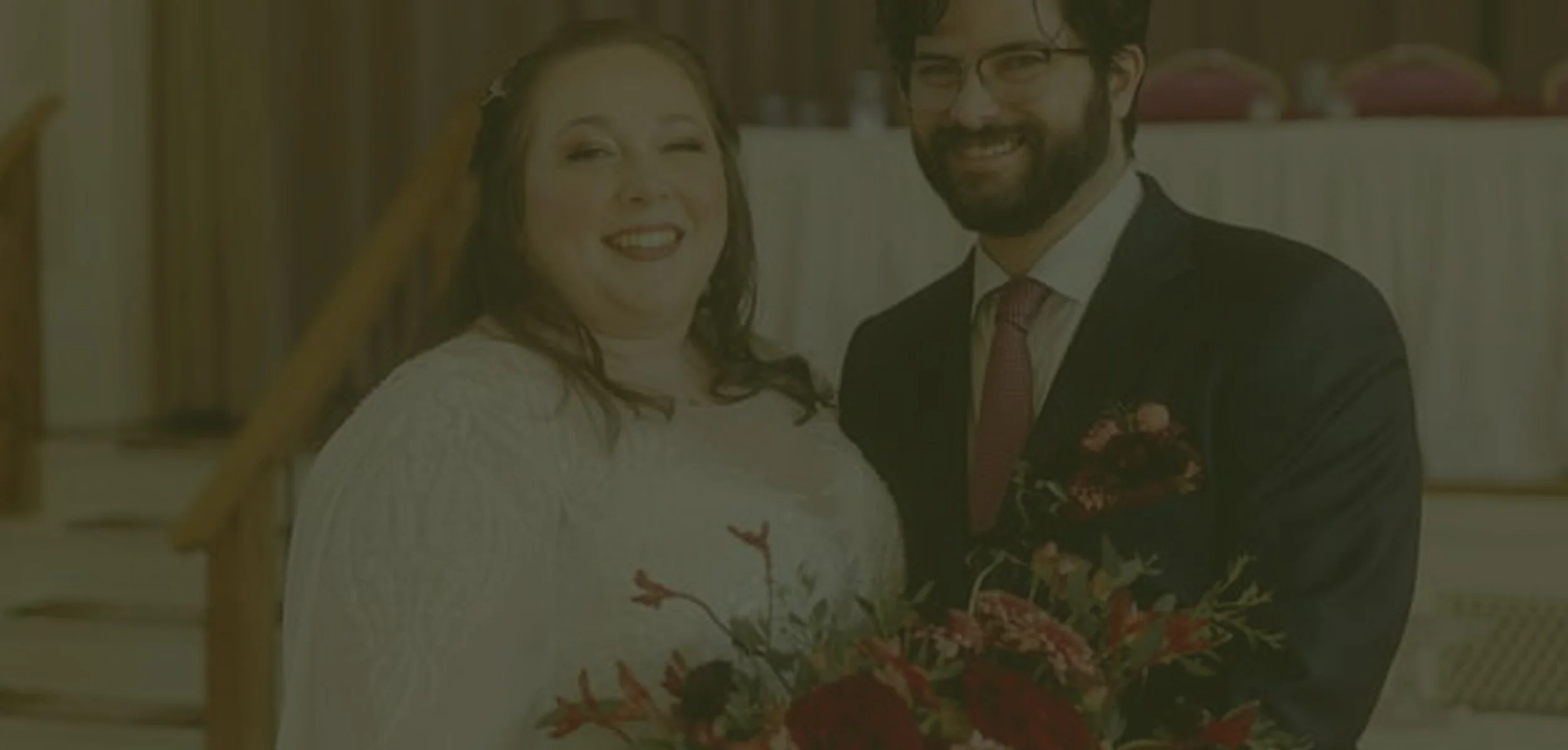 A happy woman in a white dress and a man in a black suit and tie, both smiling, standing close together with the woman holding a bouquet of flowers in an indoor setting.
