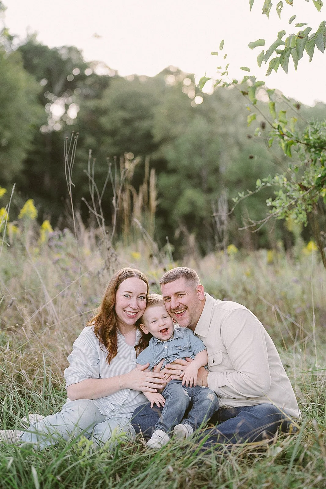 A happy family of three, a woman, a man, and a young boy, sitting on the grass in a natural outdoor setting with trees, smiling and enjoying each other's company.