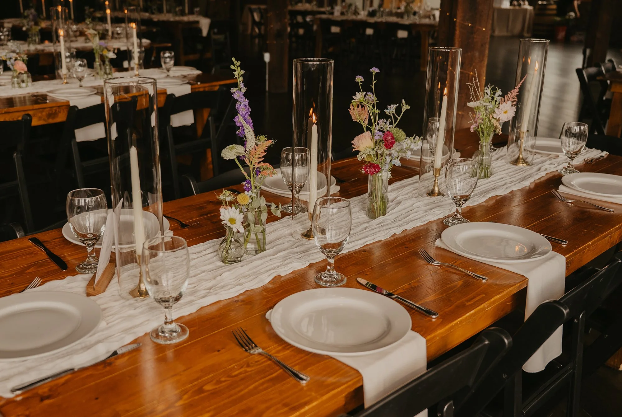 Wedding table decorated with white runner, white plates, silverware, wine glasses, tall clear vases with candles inside, and smaller vases with assorted flowers, in a rustic venue.