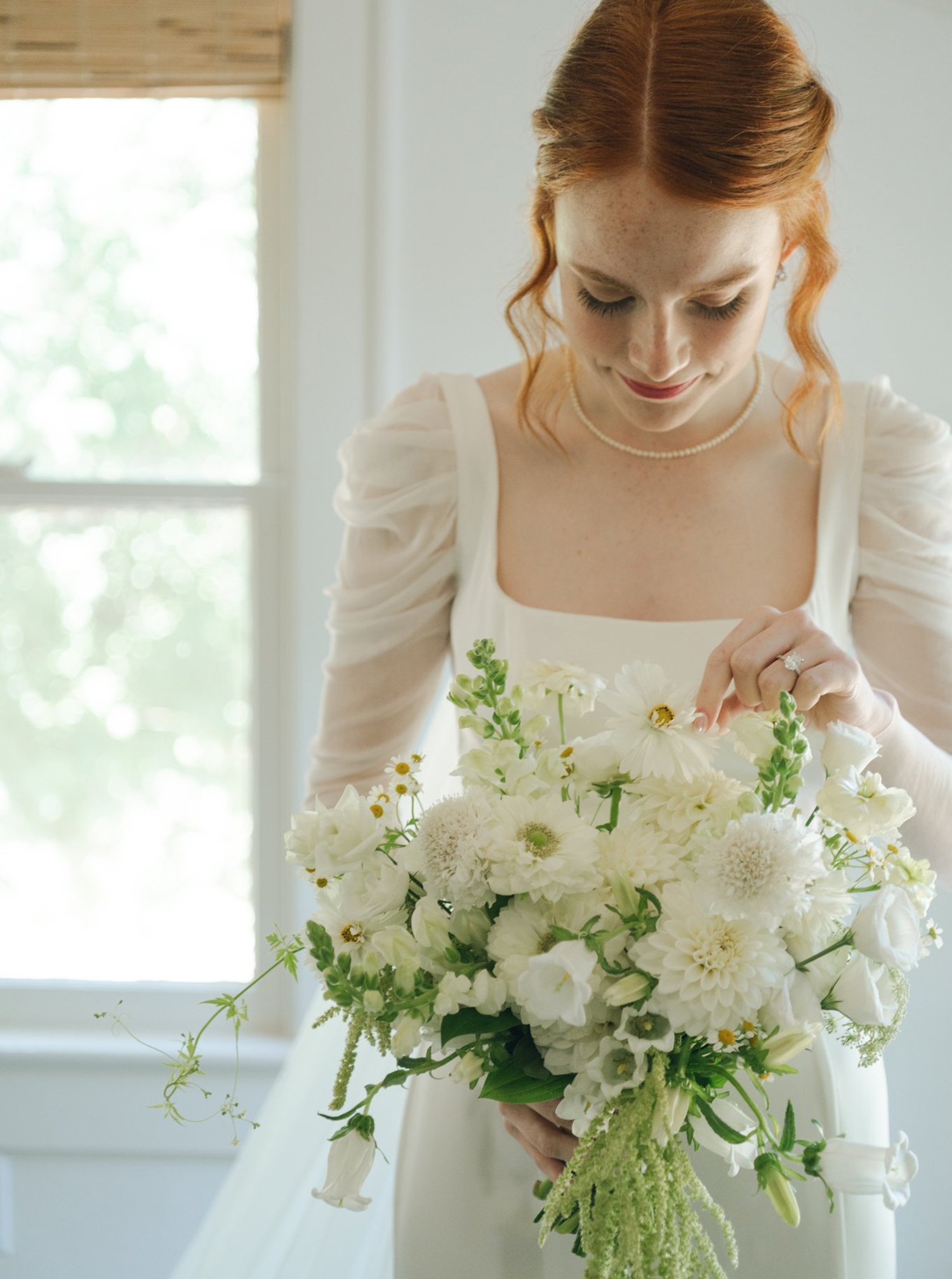 A bride with red hair in a white wedding dress holding a bouquet of white flowers, standing near a window.