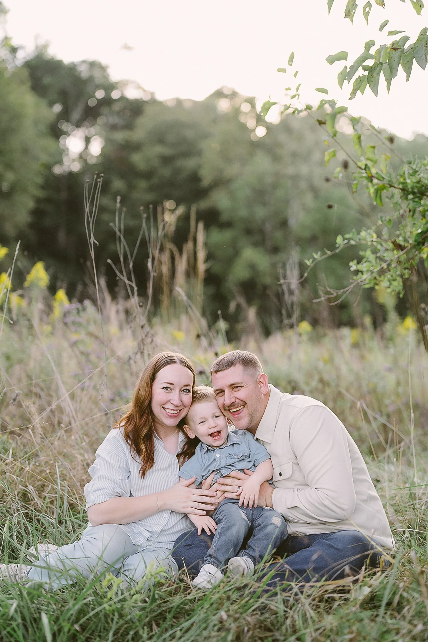 A happy family of three sitting on grass in a field with trees in the background during sunset, smiling and embracing.