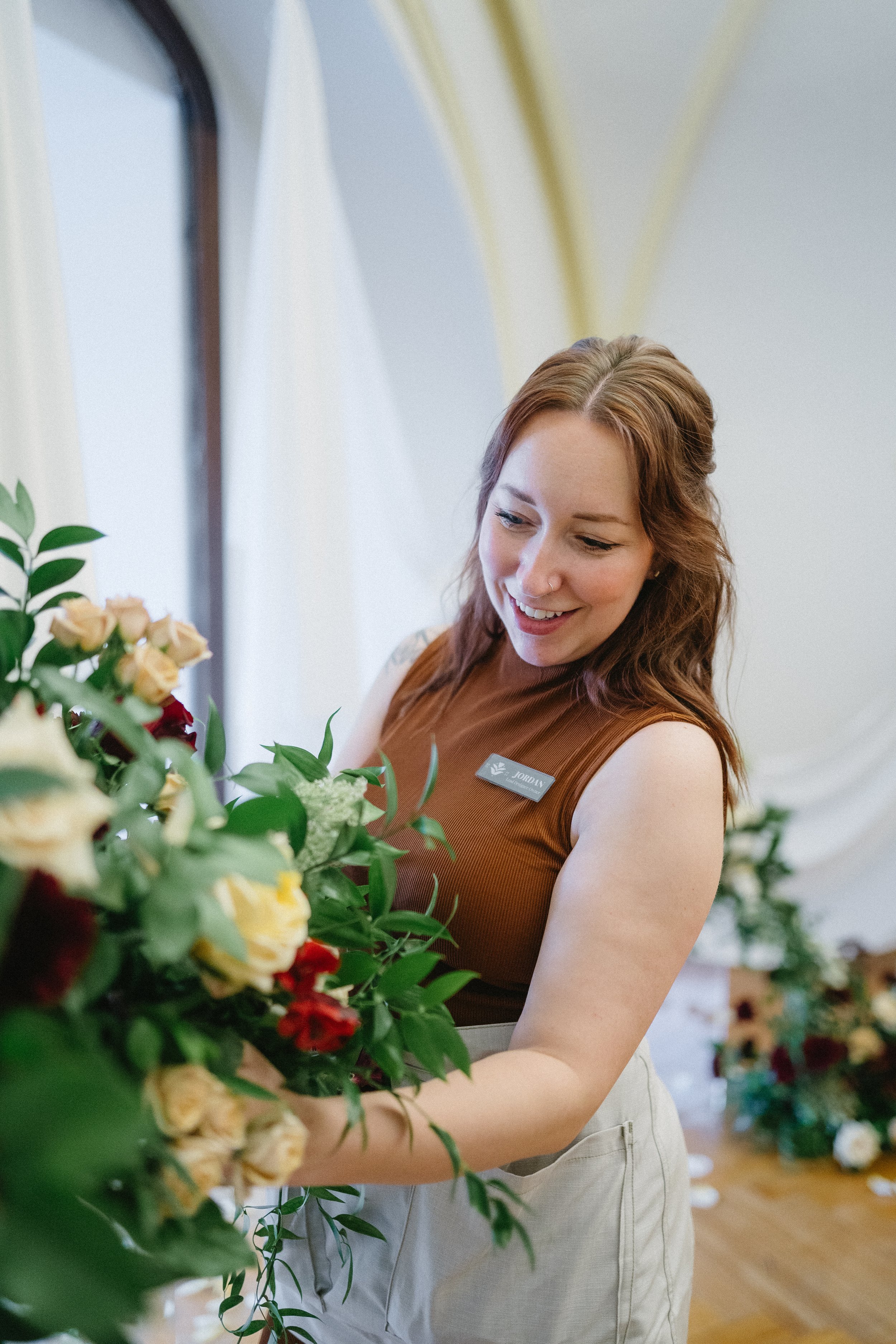 A woman with red hair and a name tag arranging a bouquet of flowers in a bright room with draped white curtains.