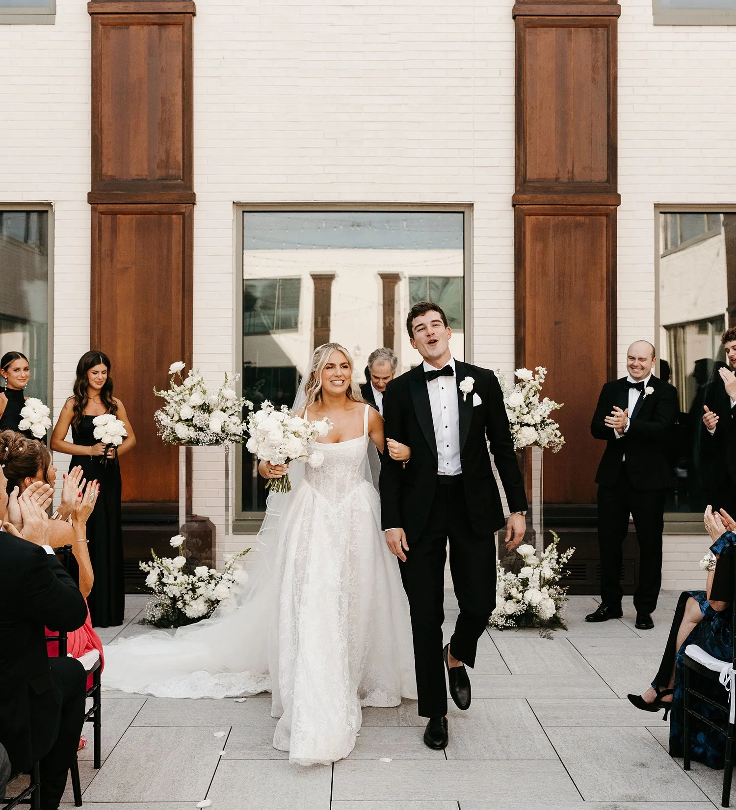 Bridal couple walking down the aisle, bride in a white wedding dress holding a custom floral bouquet and groom in a tuxedo, with guests applauding on either side.