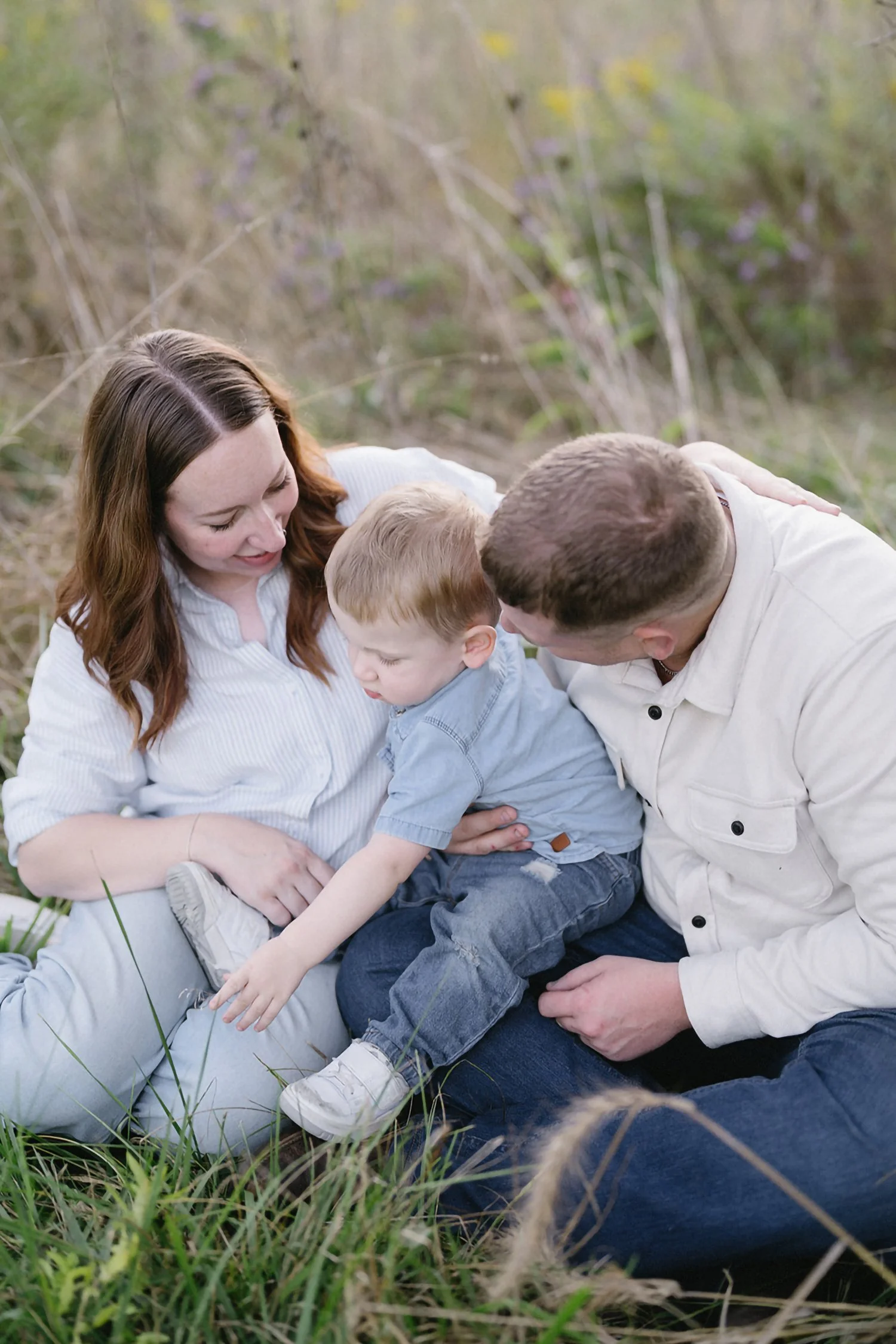 A family with a woman, a man, and a young boy sitting on the grass outdoors, surrounded by tall grass and plants, enjoying a moment together.