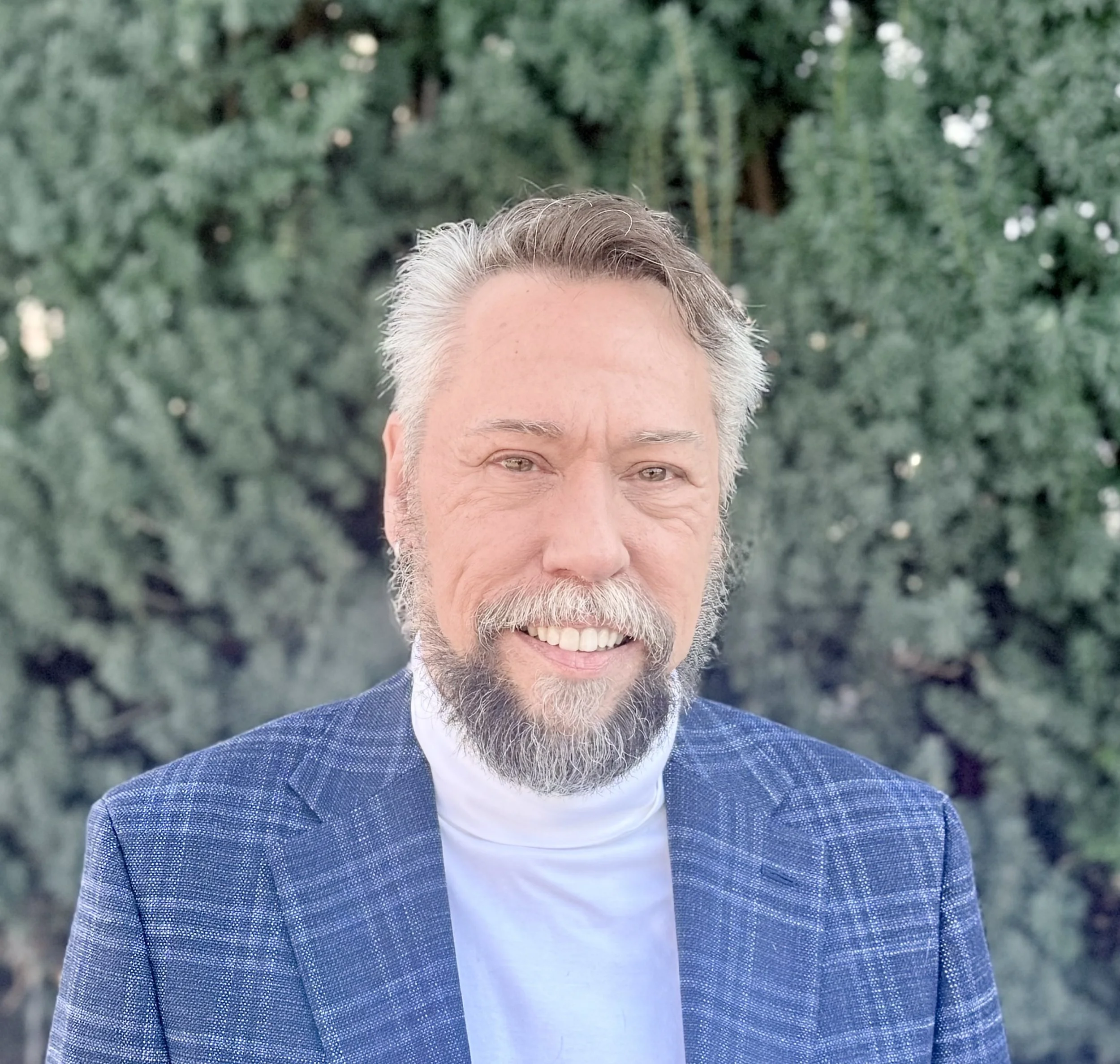 Headshot of Robb in a white turtleneck, blue tartan blazer, behind some greenery