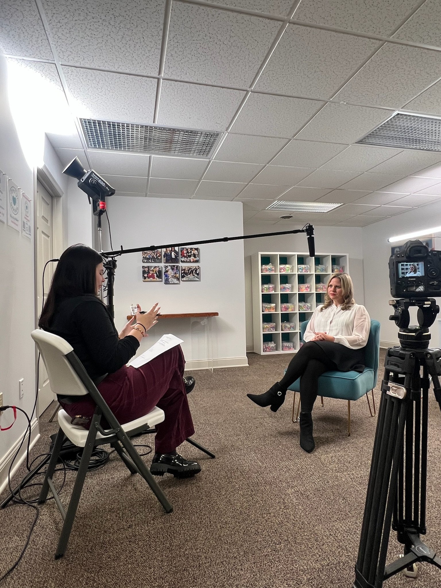 A woman is being interviewed on camera in an office or studio setting, seated on a teal chair, with a woman interviewer sitting on a white chair facing her. The interviewer has long black hair and is holding papers, a microphone boom is overhead, and there is a camera on a tripod recording the interview. The background includes a shelving unit with small containers and photos on the wall.