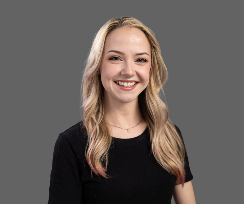A young woman with long blonde hair, smiling, wearing a black shirt and a delicate necklace, against a plain gray background.