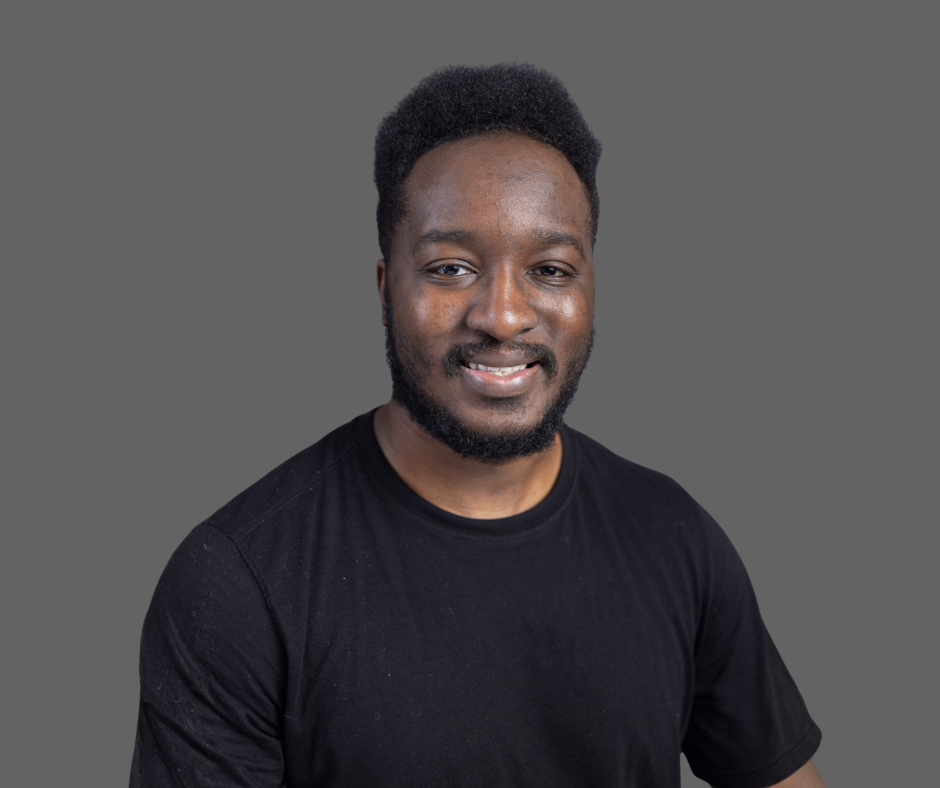 A young man with dark skin, short curly hair, and a beard, wearing a black t-shirt, smiling at the camera against a gray background.