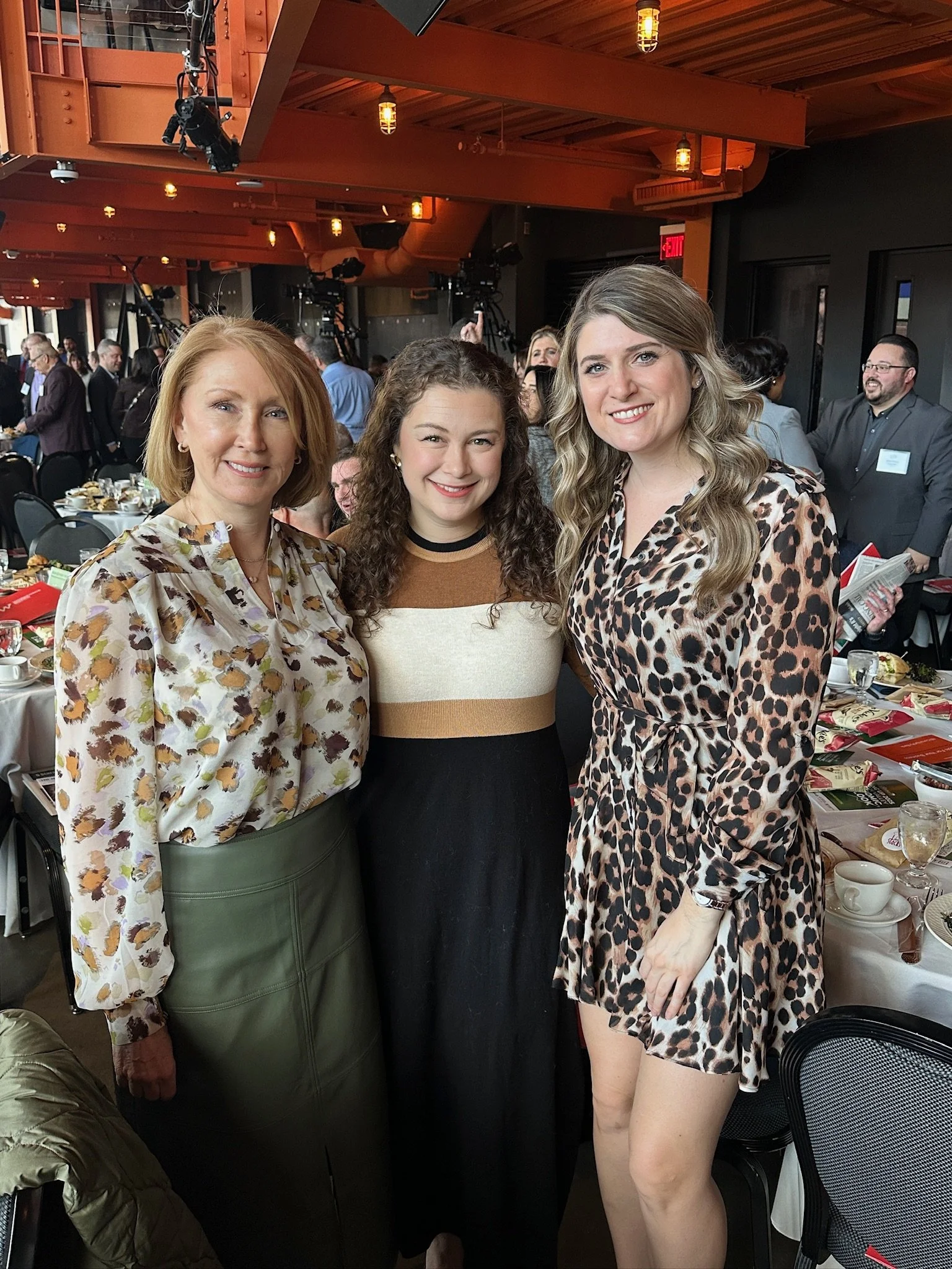 Three women standing together at a banquet or conference event, smiling at the camera in a decorated indoor venue with round tables, plates, glasses, and other guests in the background.