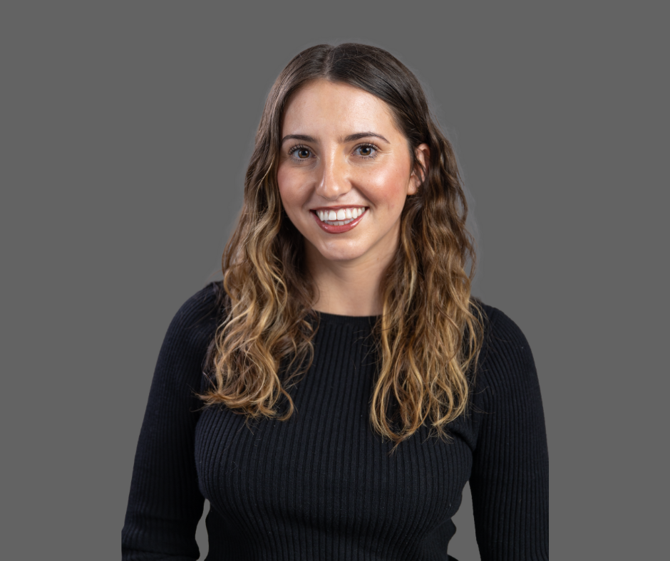 Portrait of a young woman with long, wavy hair wearing a black ribbed sweater, smiling against a gray background.