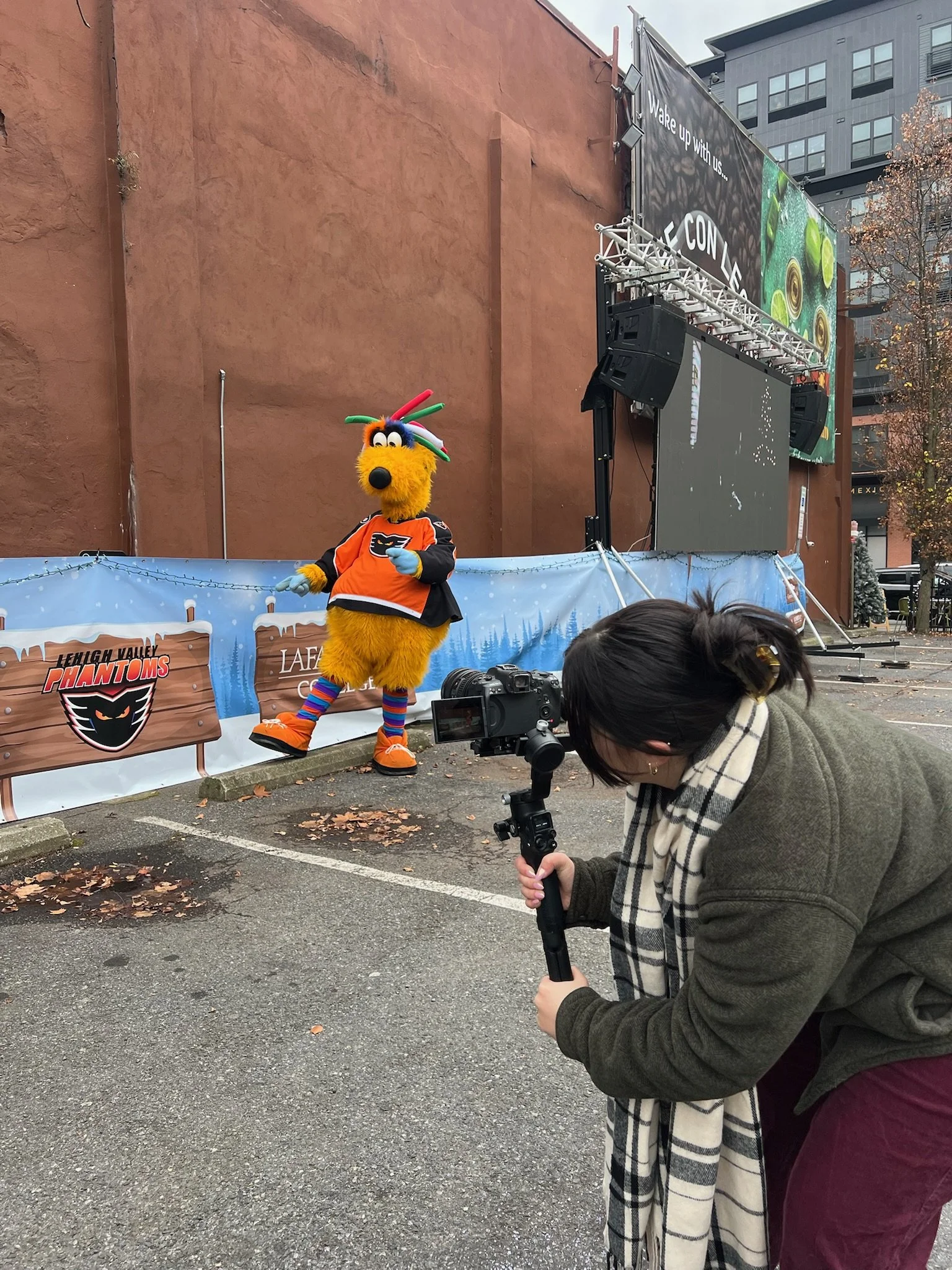 A person dressed in a brown jacket and plaid scarf is taking a photo or filming a mascot of a yellow furry animal in a sports jersey outdoors near a stage with speakers and a large screen. The mascot has a colorful balloon hat and striped socks, and is standing in front of a themed banner for the Lehigh Valley Phantoms hockey team.