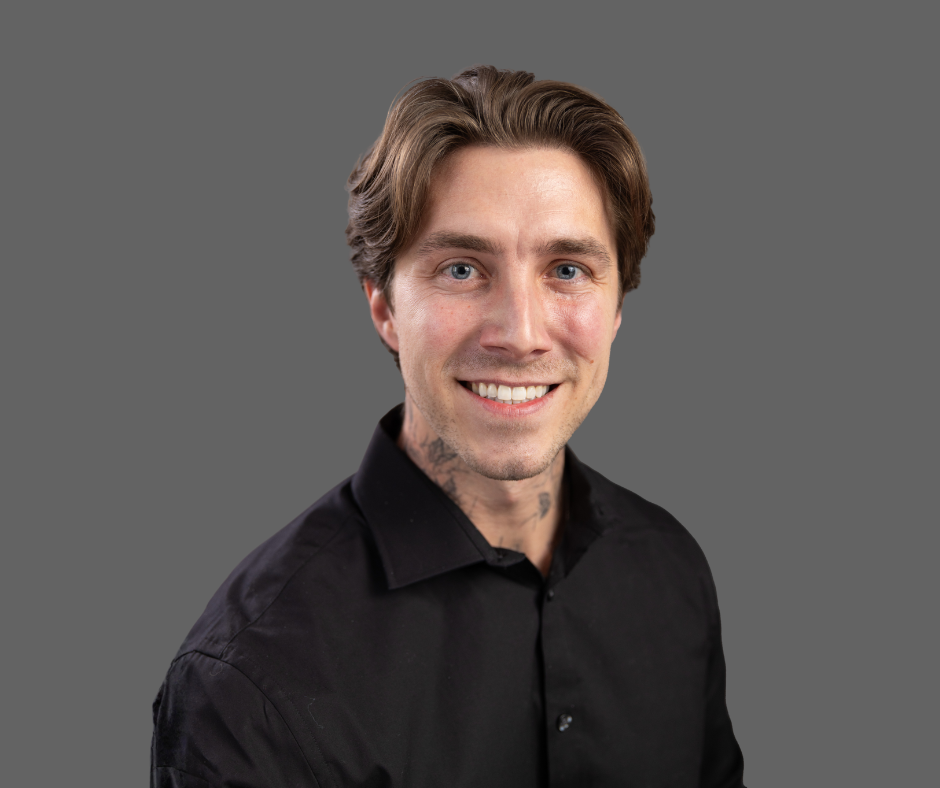 A young man with light skin, brown hair, and blue eyes smiling, wearing a black shirt, against a neutral gray background.