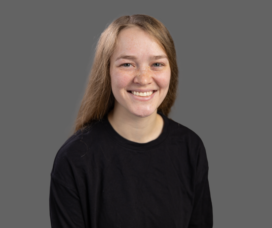 A young woman with long red hair, freckles, and blue eyes, smiling, wearing a black shirt, against a gray background.