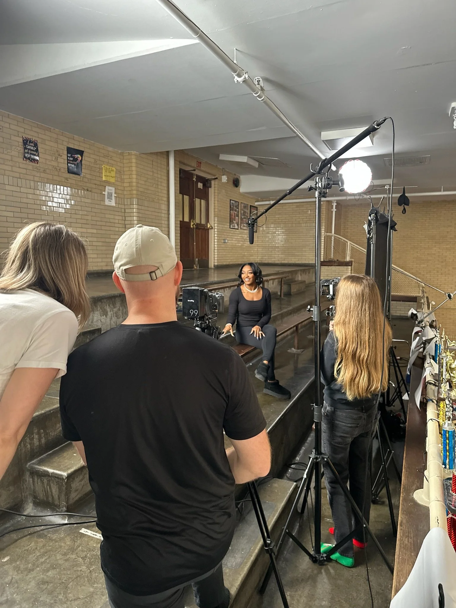 A woman sits on a step in front of a brick wall, during a filming session with multiple crew members and lighting equipment.
