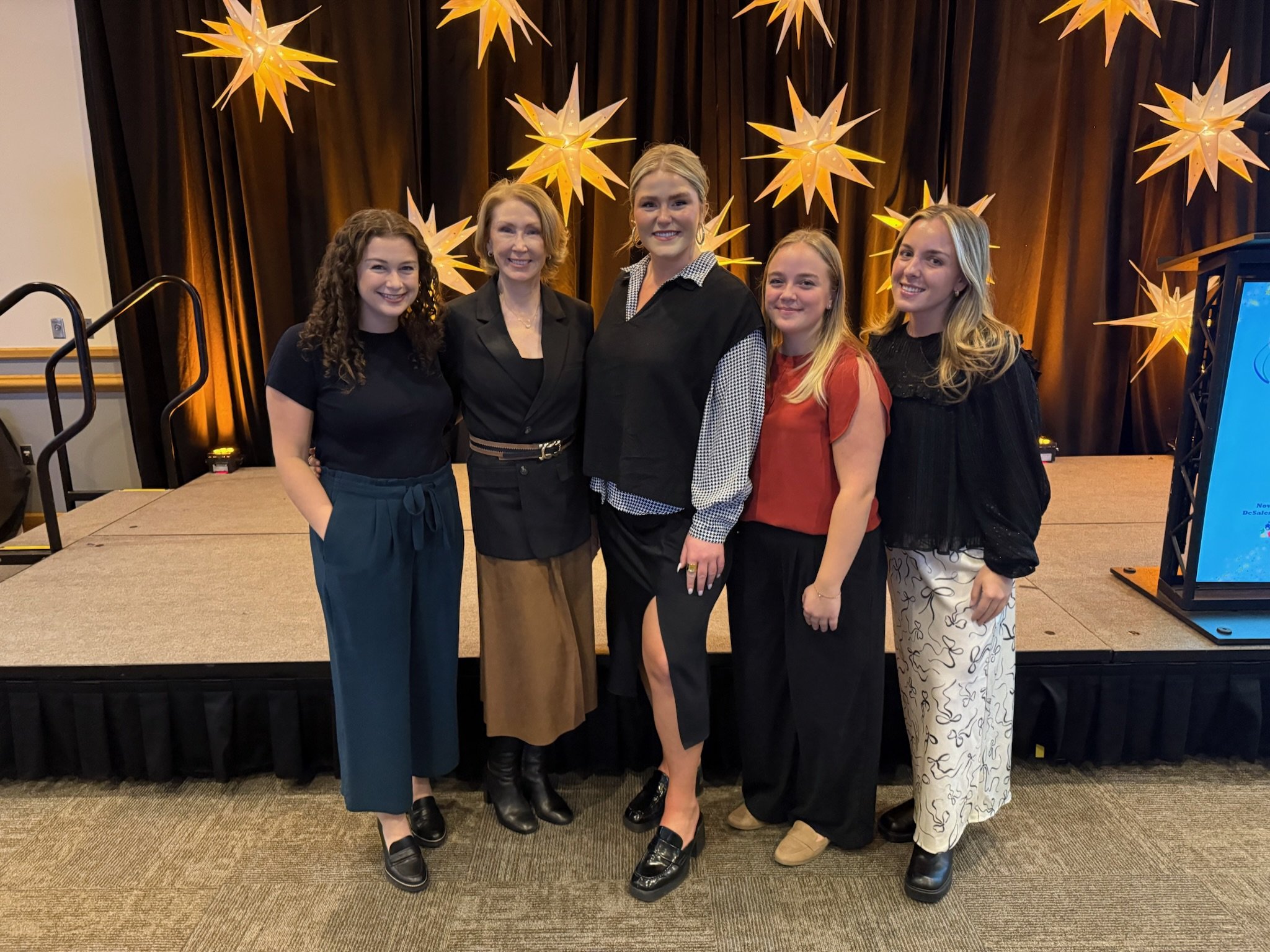 A group of five women standing on a stage in front of a dark curtain decorated with illuminated star-shaped lights, all smiling at the camera.