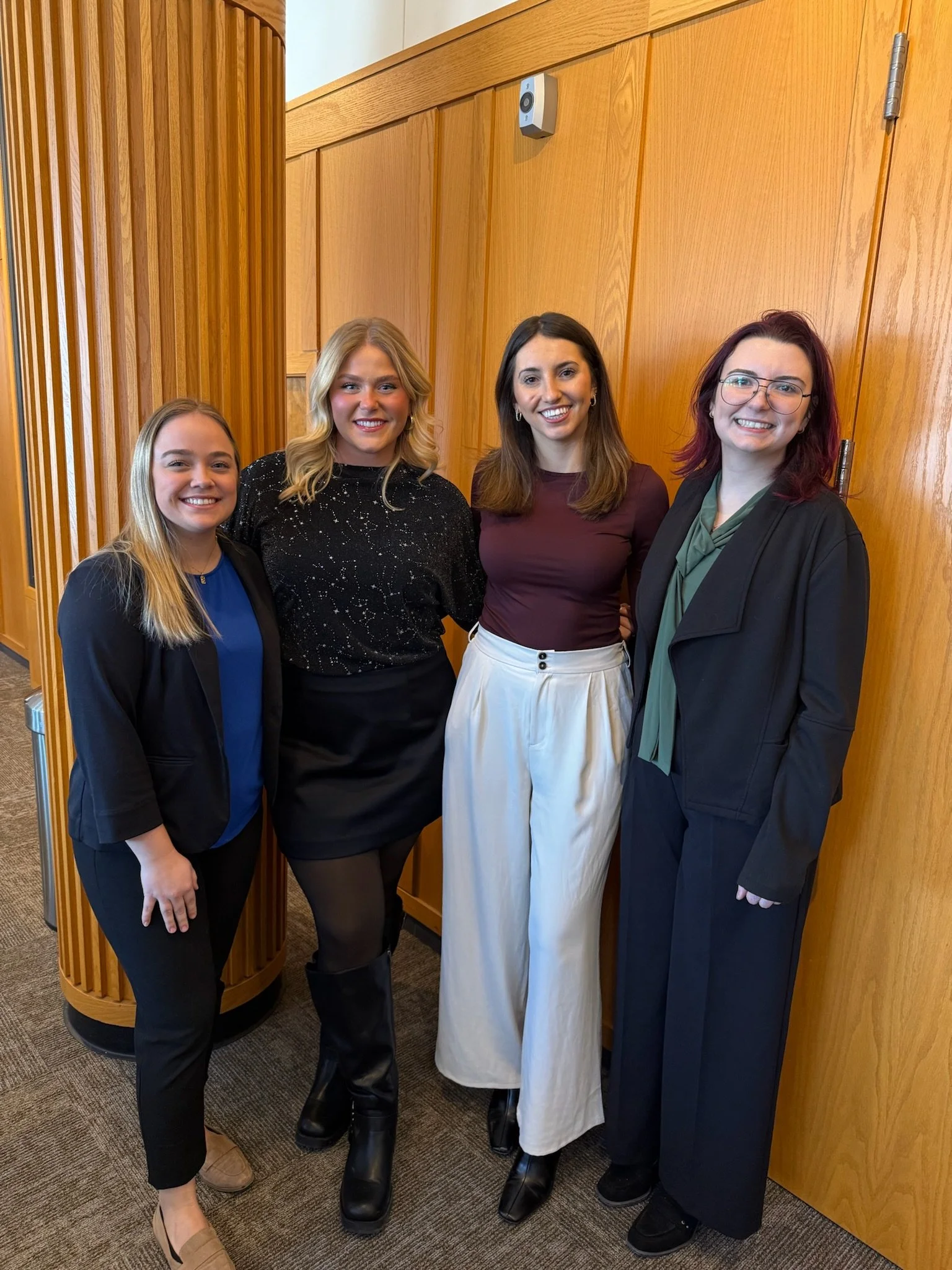 Four women standing together in front of a wooden wall, smiling at the camera.