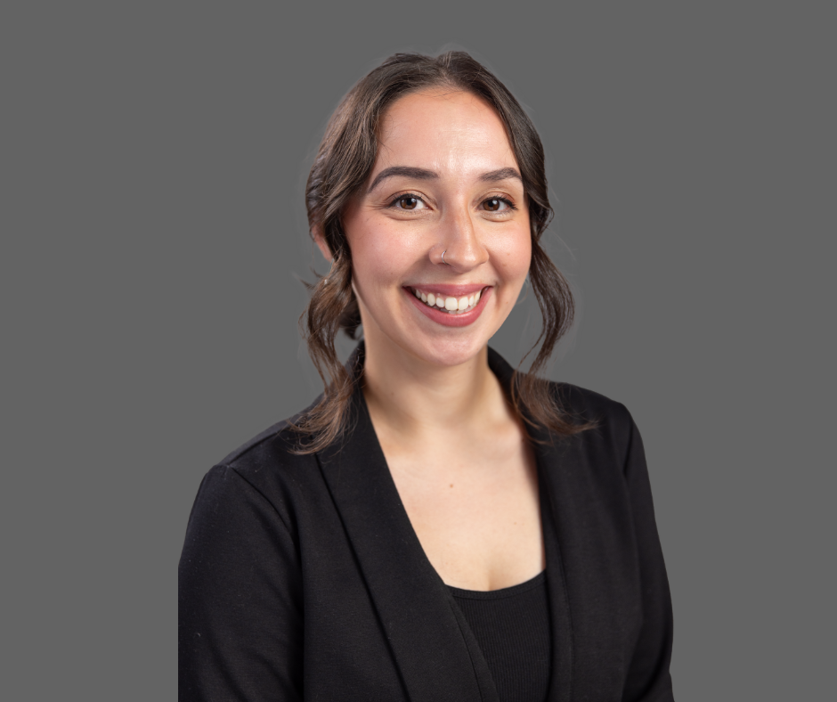 A woman smiling with dark hair, wearing a black blazer and a nose ring, against a plain gray background.