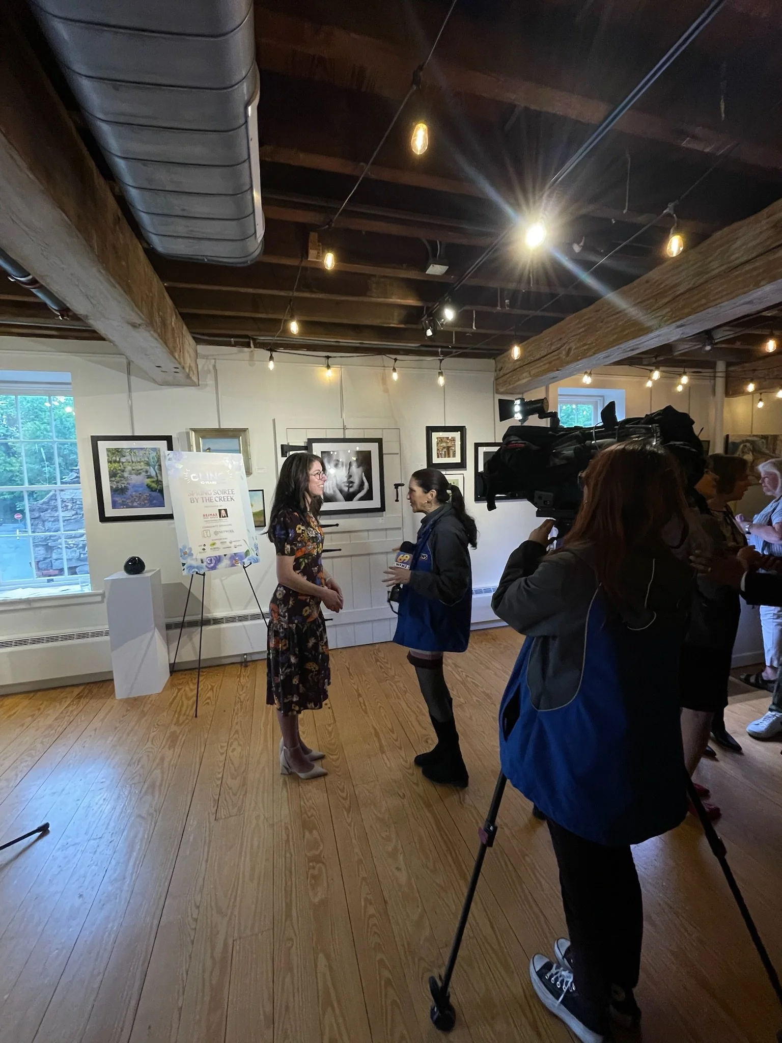 Two women interview each other in an art gallery with framed photographs on the wall. A camera operator records the interview, with safety shoes and a broadcast camera, as other people observe.
