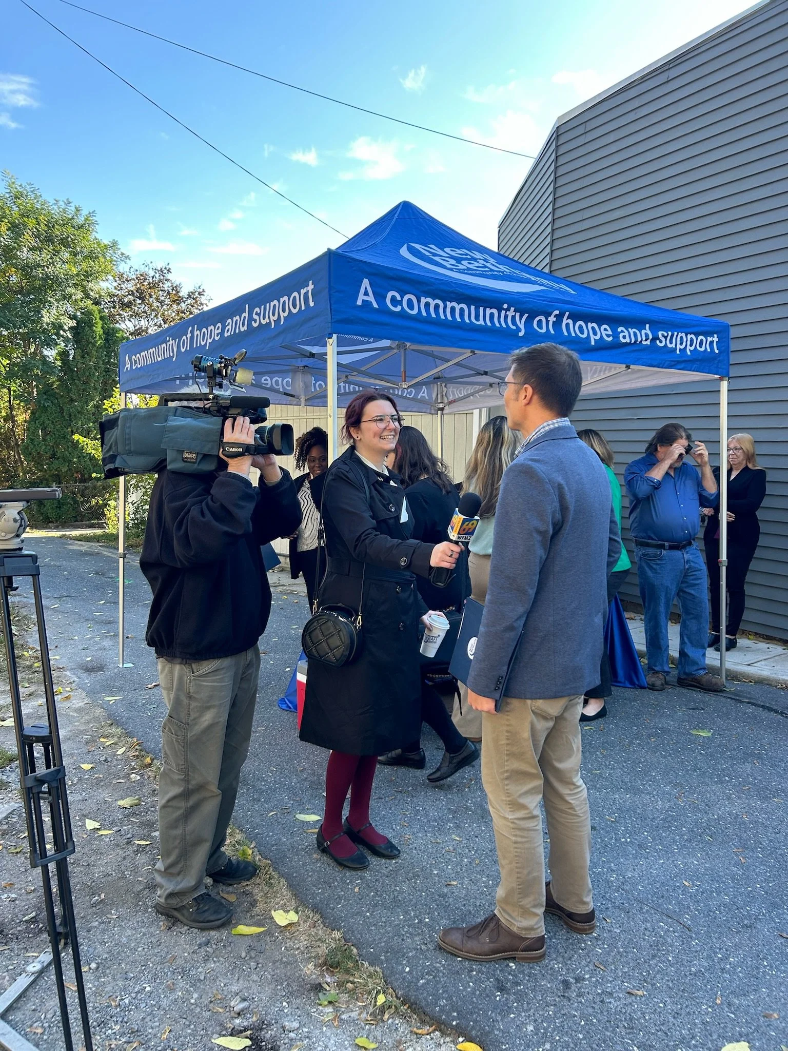 A news interview at an outdoor event with a woman reporter holding a microphone interviewing a man in a blue blazer near a blue canopy tent that reads 'A community of hope and support.'