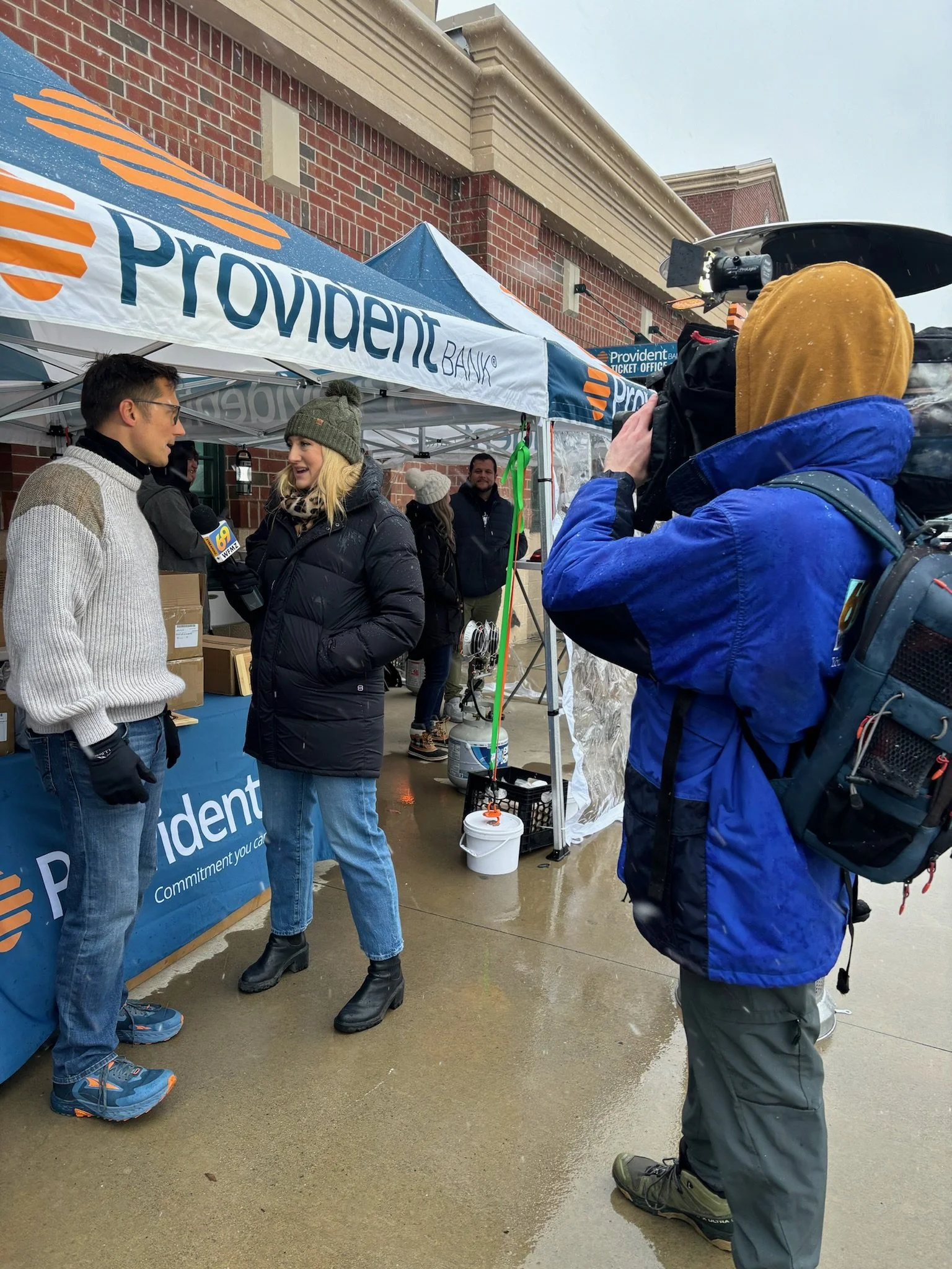 A television news crew interviews a woman at an outdoor Providence Bank event booth on a rainy day. The woman is wearing a gray beanie and black coat, while the man being interviewed wears glasses and a gray sweater. The camera operator is dressed in a blue jacket with a backpack, capturing the interview.