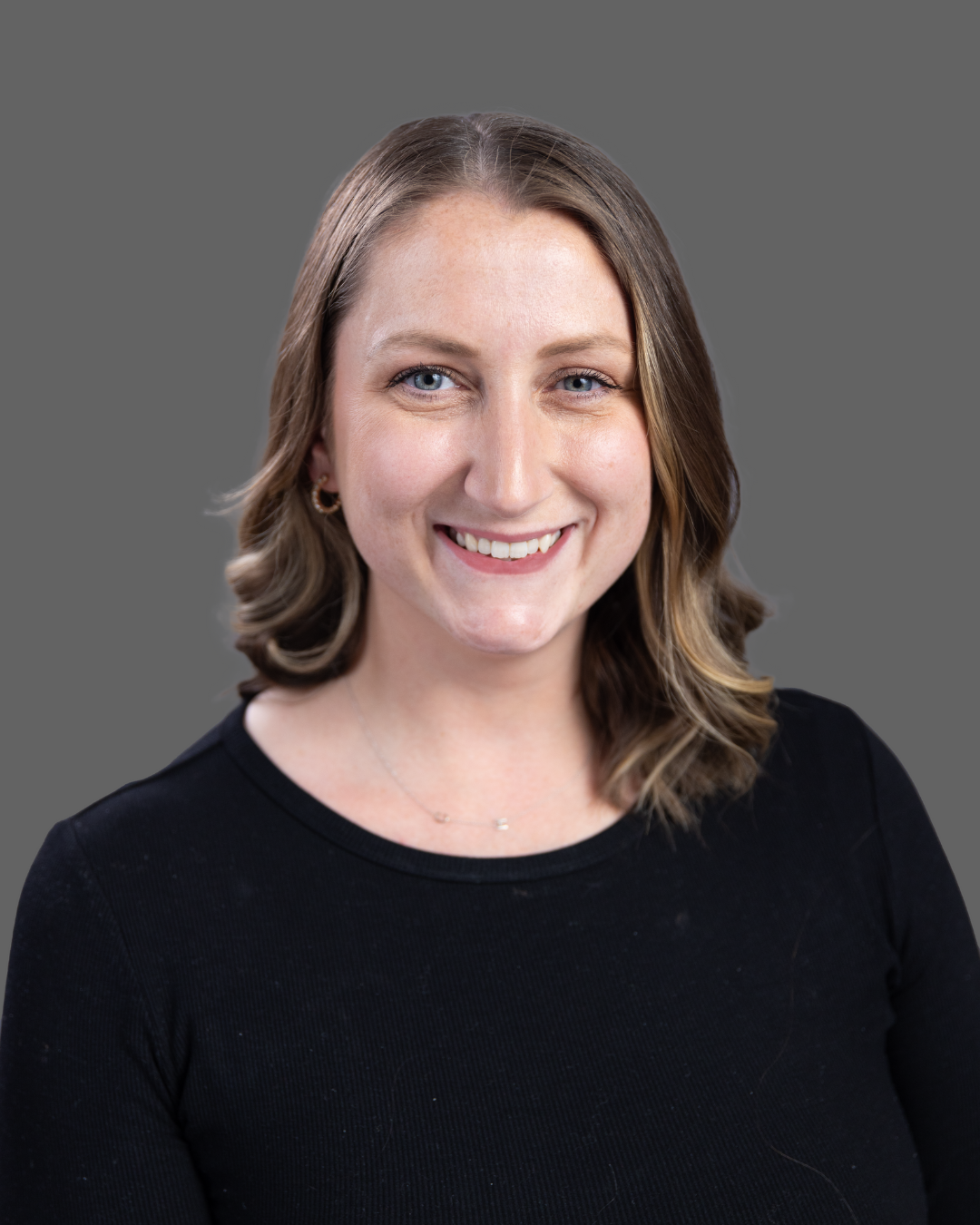 Close-up portrait of a woman with shoulder-length light brown hair, blue eyes, smiling, wearing a black top and small earrings, against a plain gray background.