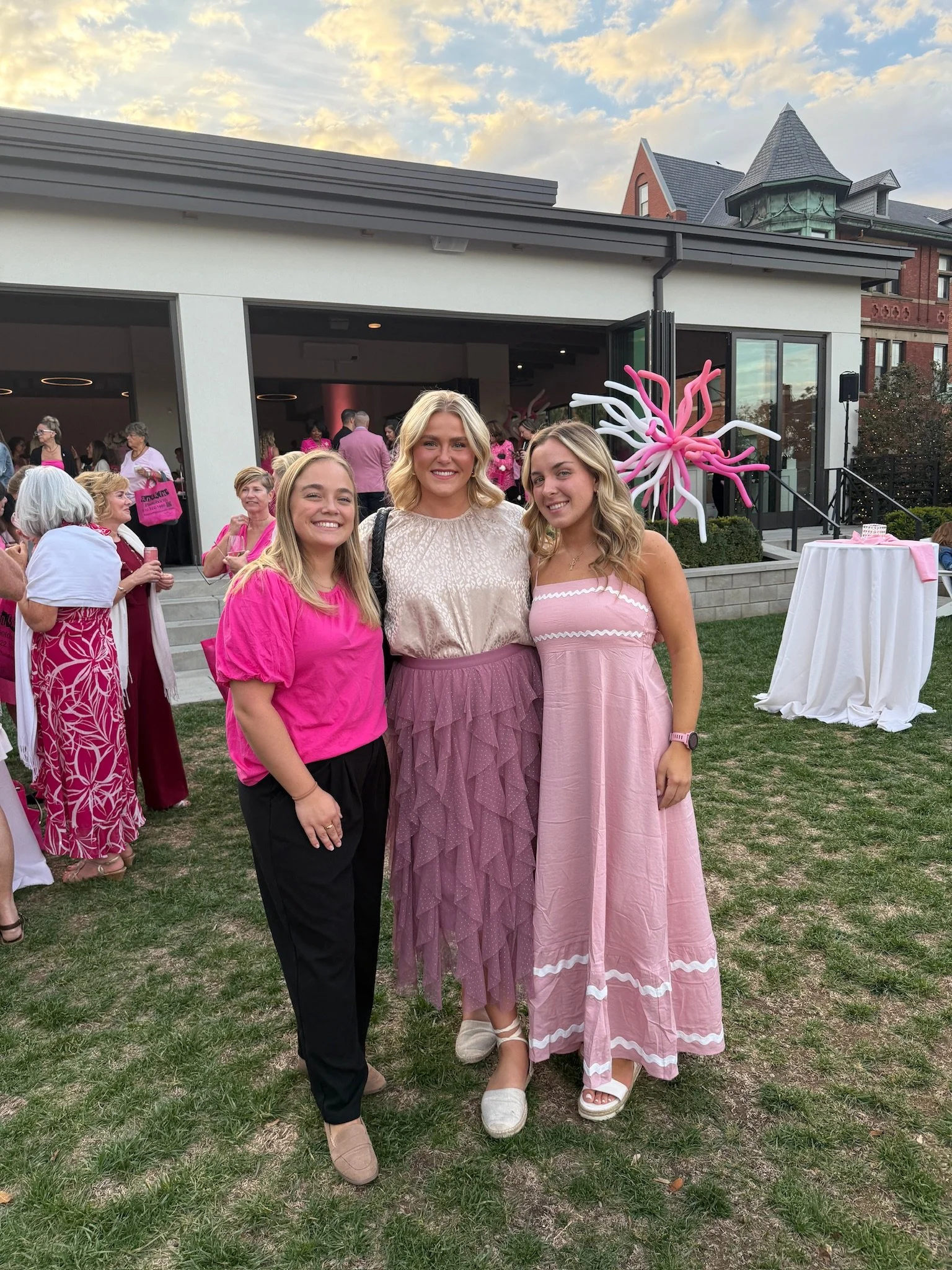 Three women at a social gathering outdoors, wearing pink and purple dresses, smiling for the camera with other guests in the background.