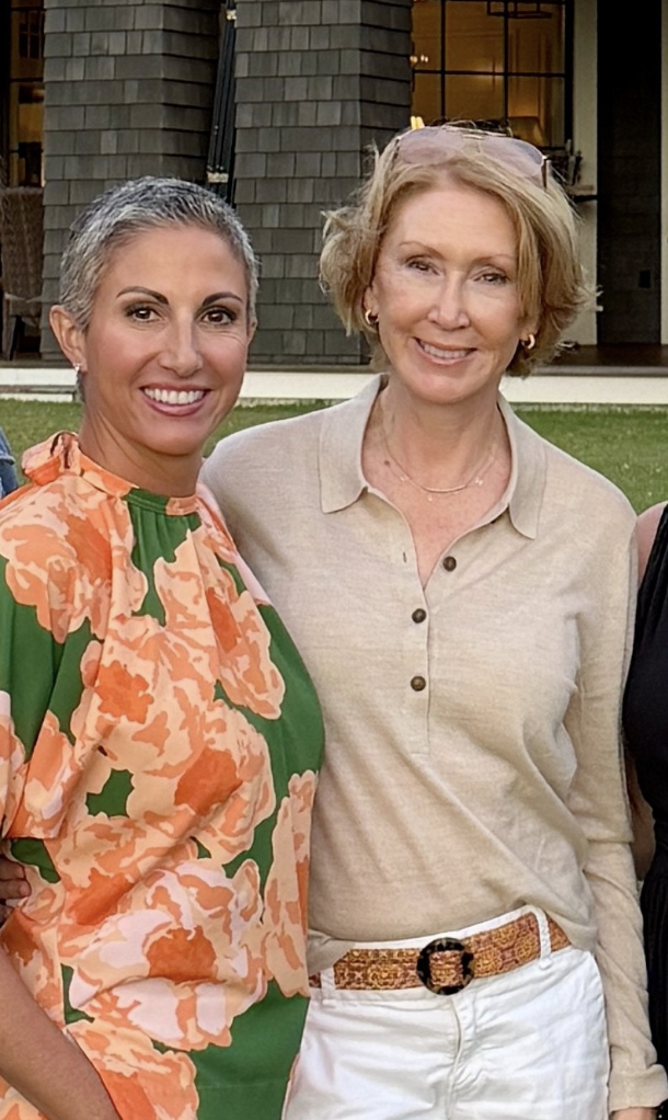 Two women smiling outdoors in front of a large building, one wearing a floral dress and the other in a beige shirt and white pants.
