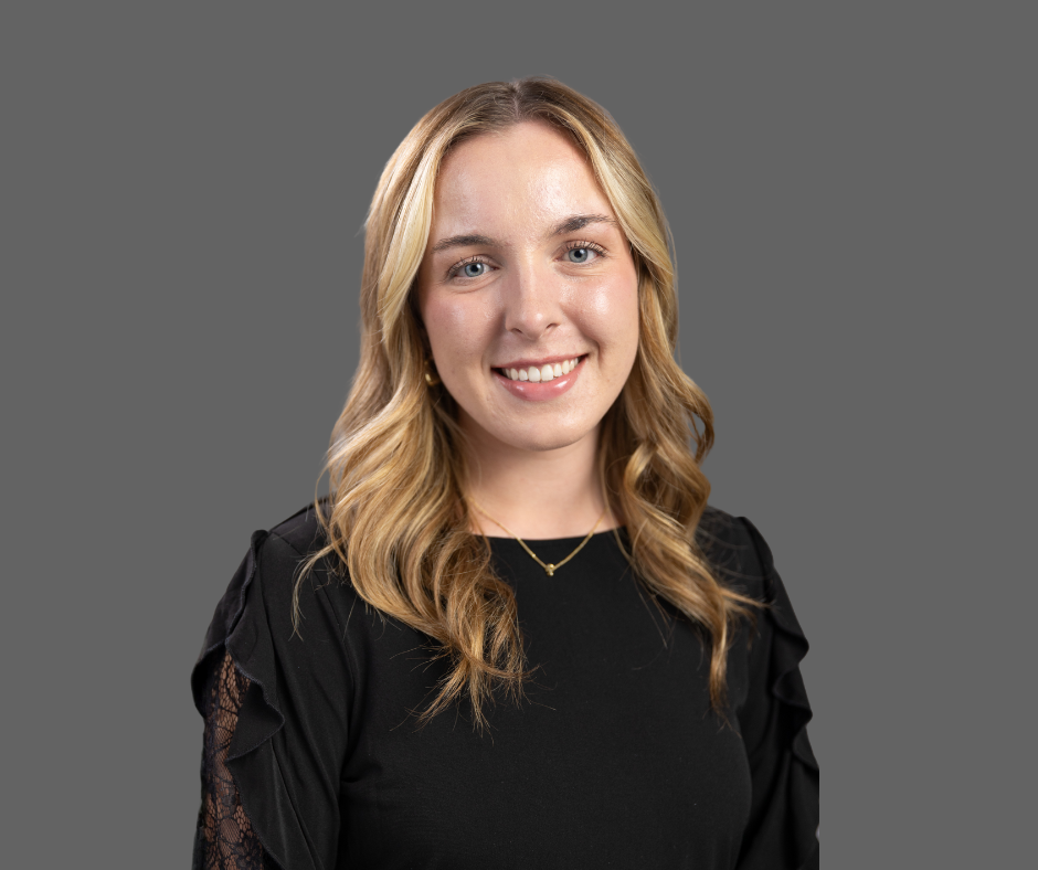 Portrait of a young woman with blonde hair, wearing a black top with lace sleeves, smiling, against a gray background.