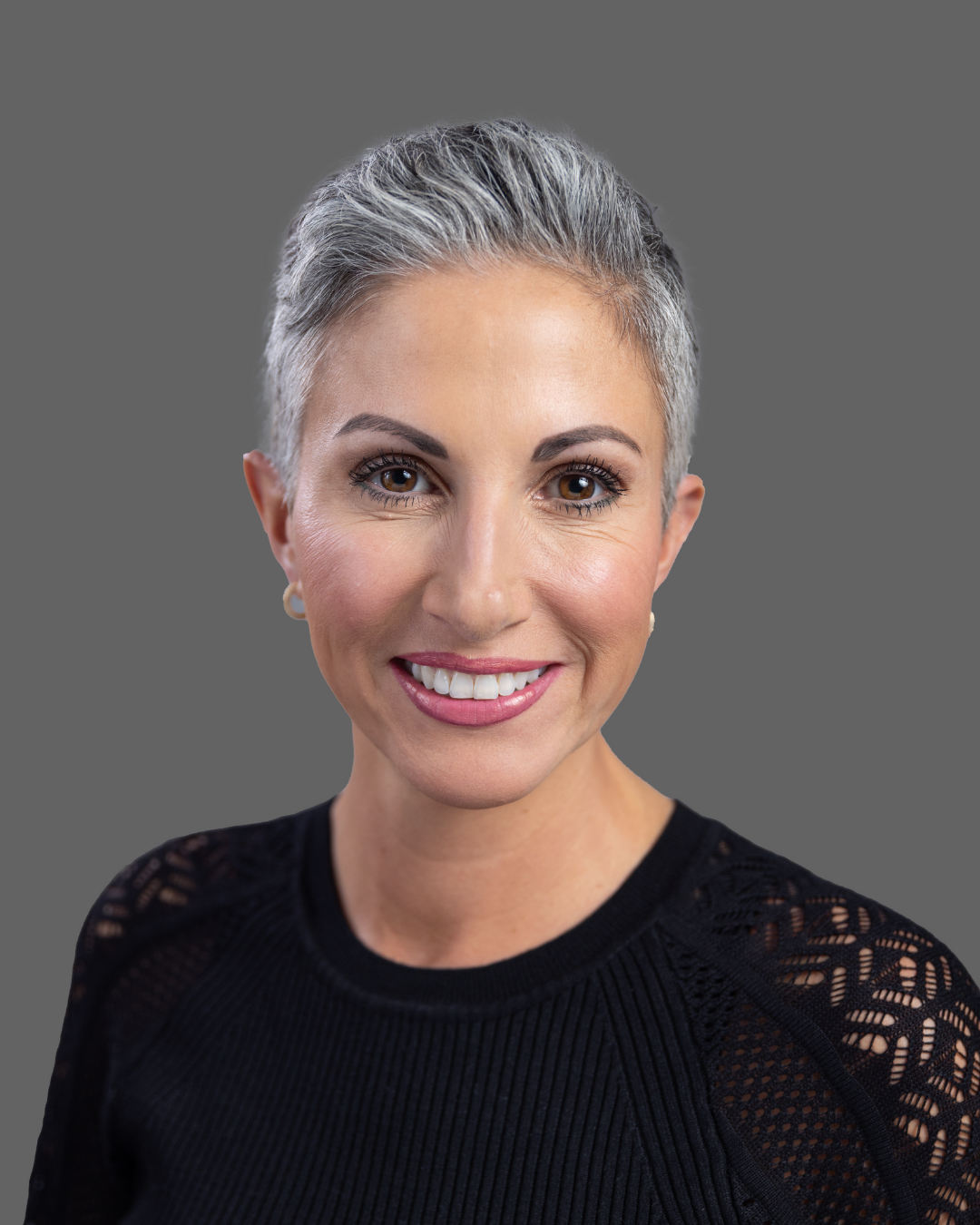 A middle-aged woman with short, gray hair and brown eyes, smiling at the camera, wearing a black textured top with lace details on the shoulders, against a gray background.