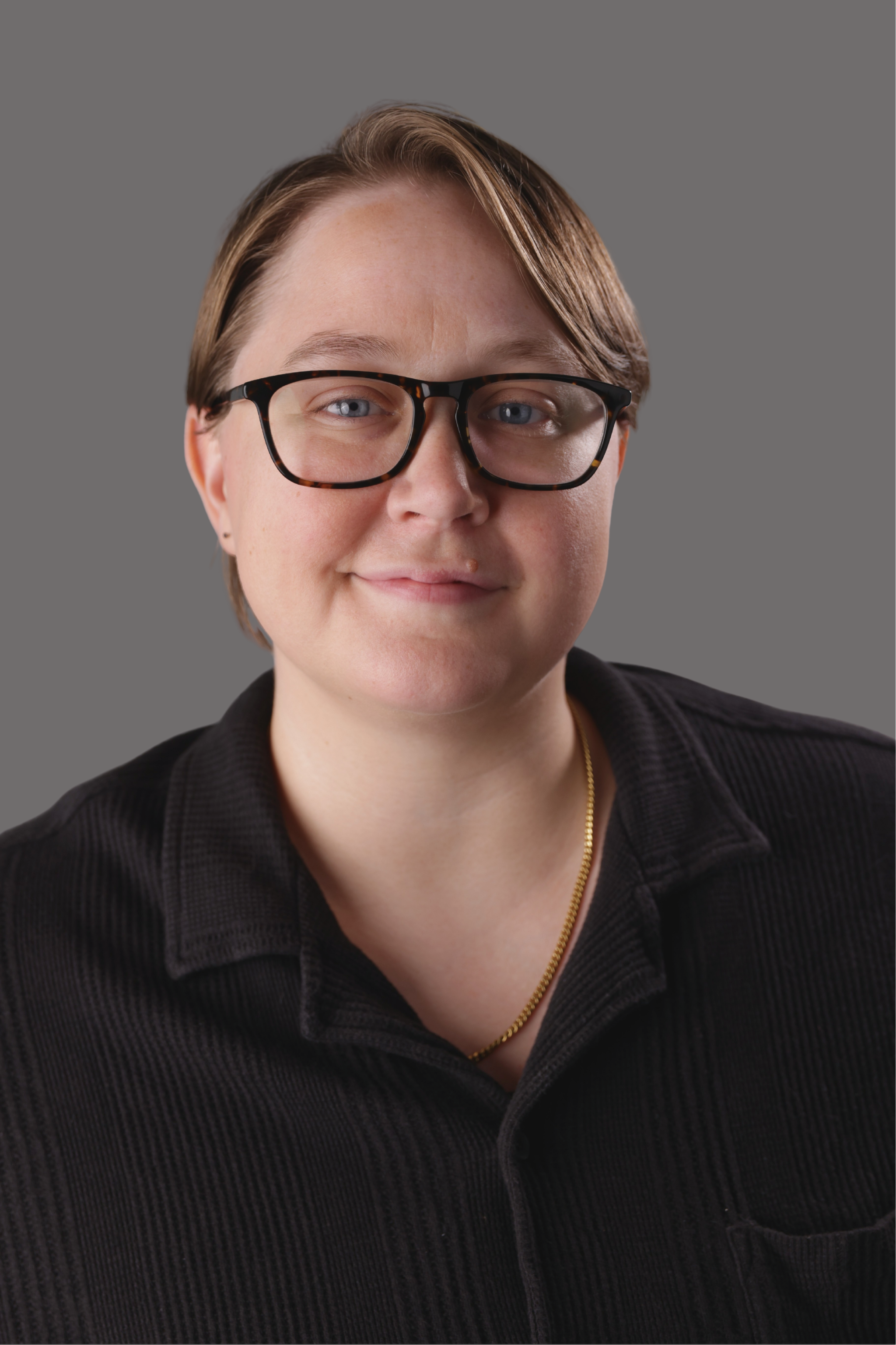 Headshot of a woman with short brown hair, wearing glasses with tortoiseshell frames, a black collared shirt, and a gold necklace, positioned against a gray background.