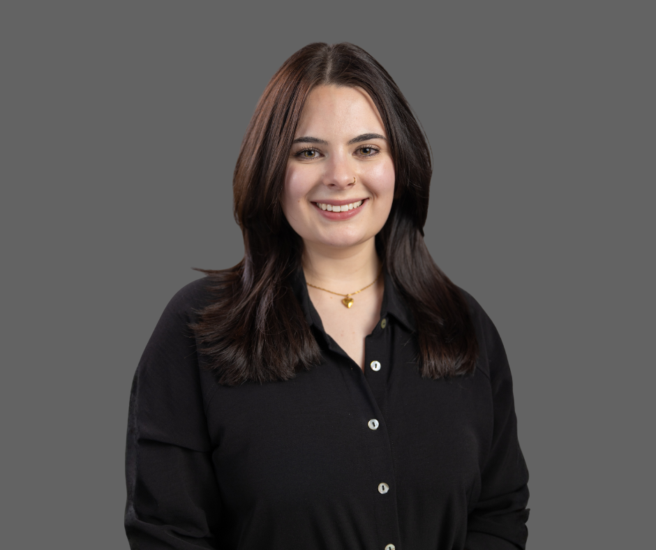 A young woman with shoulder-length brown hair, smiling, wearing a black shirt and a gold necklace with a heart pendant, standing against a gray background.