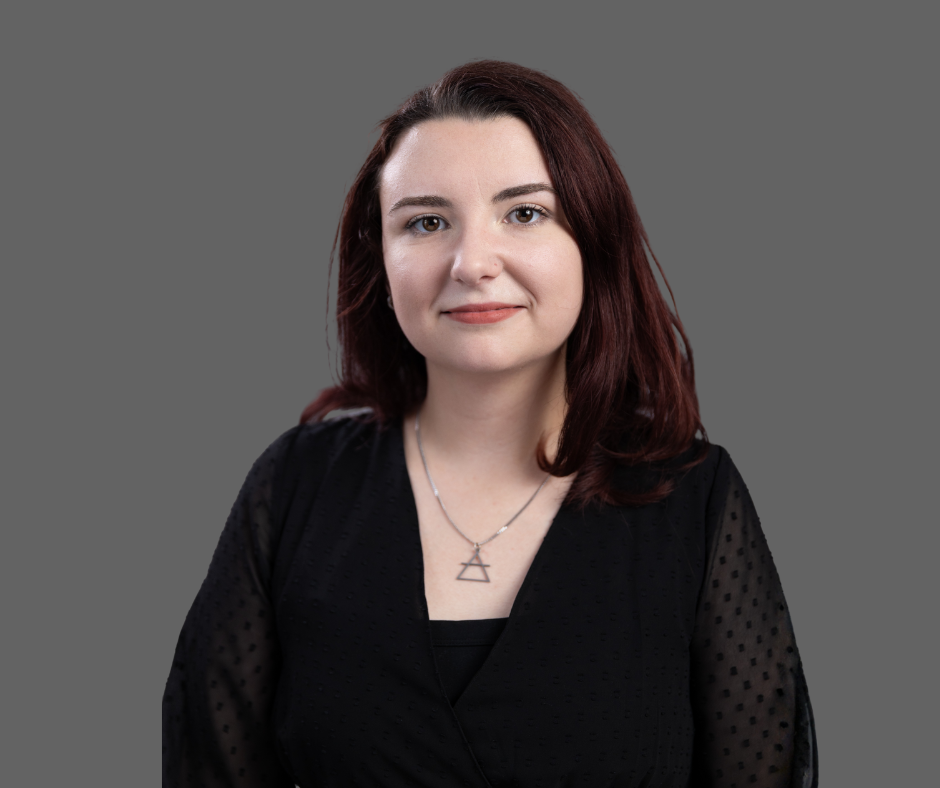 A portrait of a young woman with shoulder-length brown hair, wearing a black top with sheer sleeves, and a silver necklace with a geometric pendant, against a gray background.