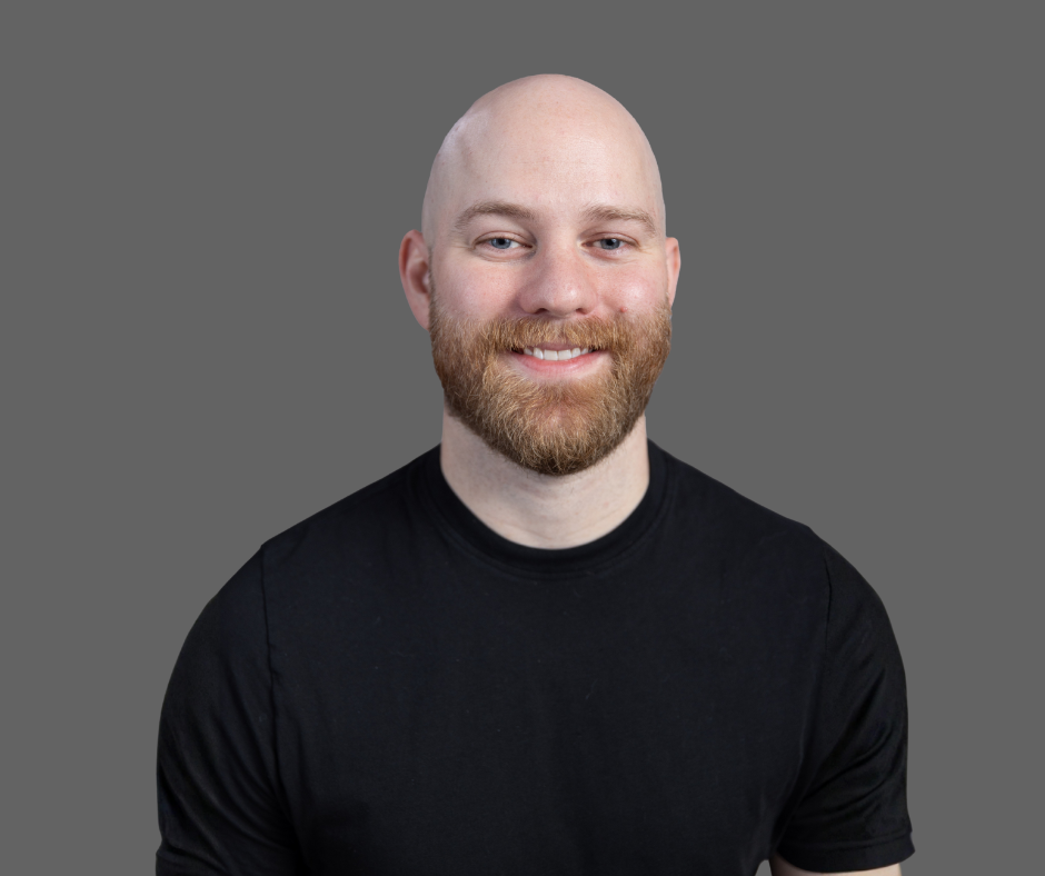 A smiling man with a beard, bald head, wearing a black t-shirt, against a gray background.
