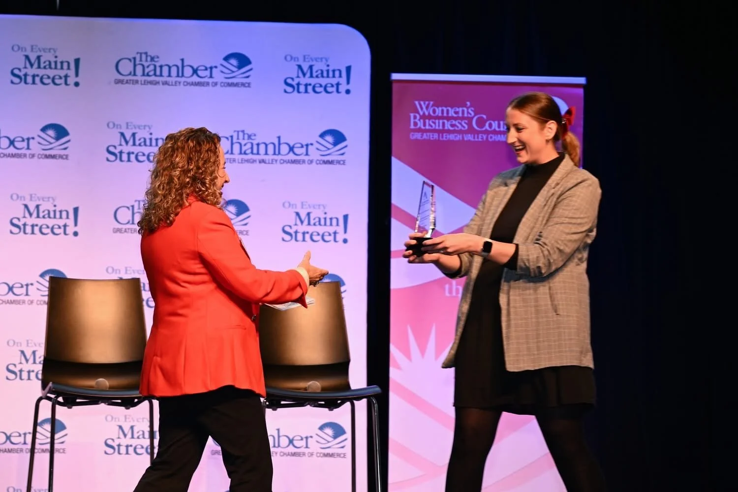 A woman in a red blazer receives an award from a woman in a plaid blazer on stage at an event hosted by the Greater Lehigh Valley Chamber of Commerce and Women's Business Council.