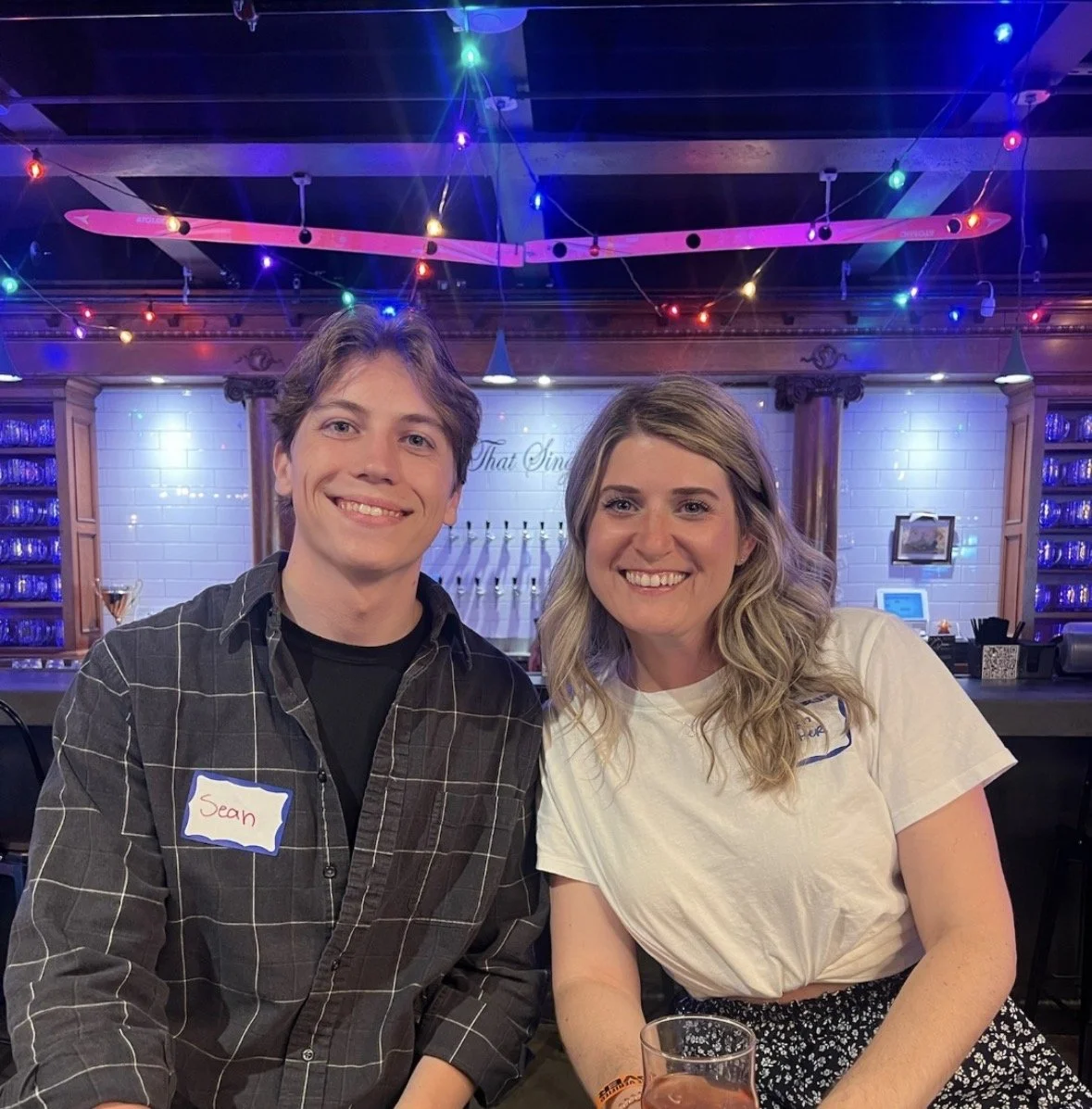 A young man and woman sitting together at a bar, smiling at the camera. The man has light brown hair and is wearing a black and gray checkered shirt with a name tag that says 'Sean'. The woman has long, blonde wavy hair and is wearing a white t-shirt. The bar has colorful string lights and a pink skateboard hanging from the ceiling.