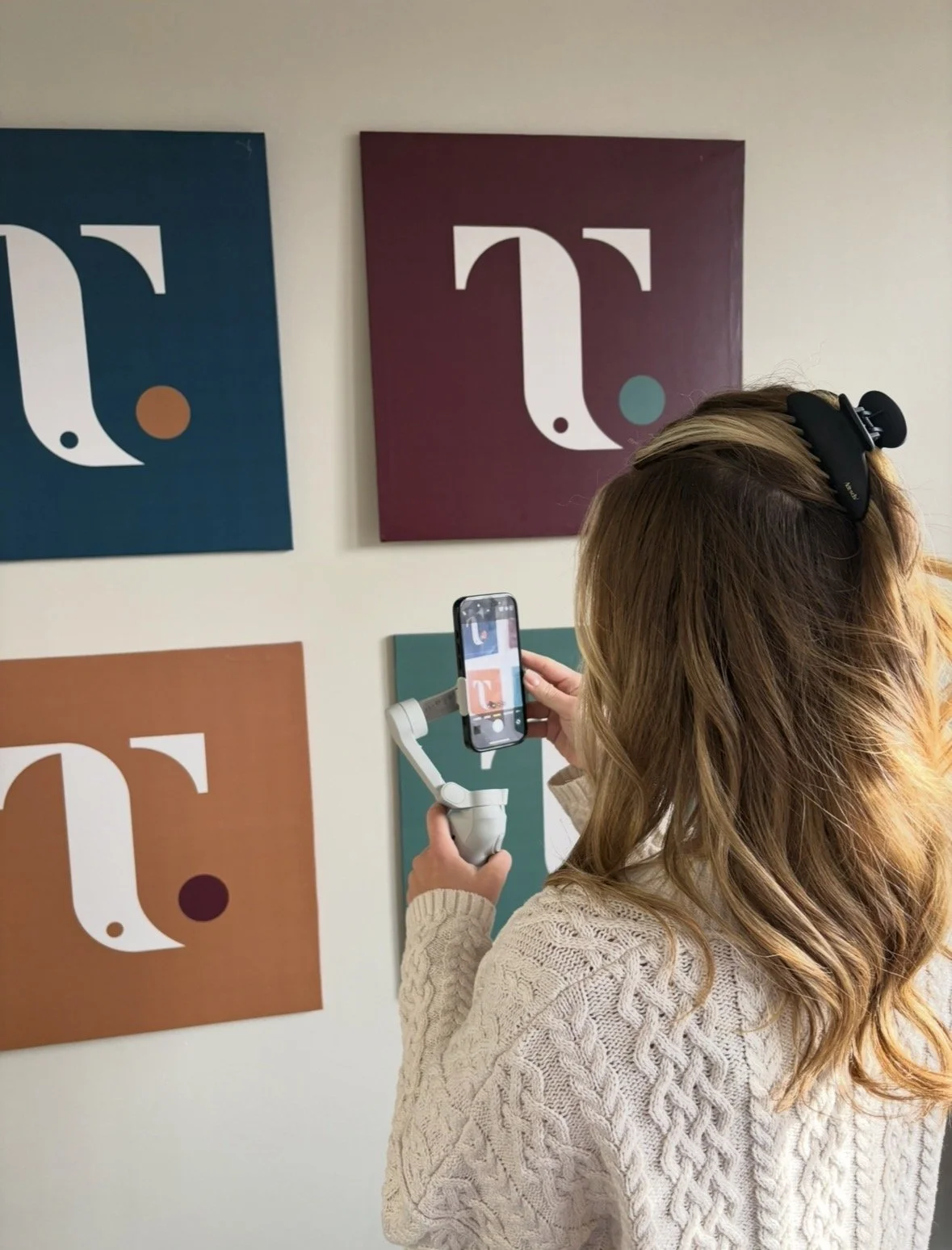A woman with brown hair wearing a cream sweater uses a smartphone mounted on a stabilizer to photograph colorful modern art on a white wall.