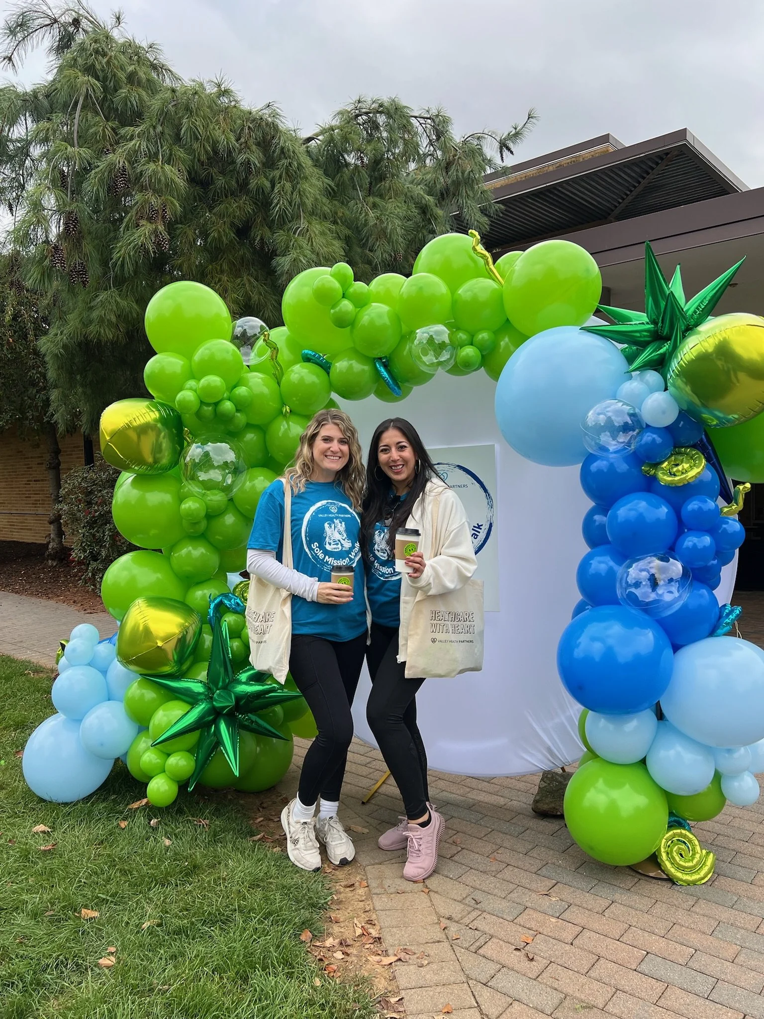 Two women standing in front of a colorful balloon arch at an outdoor event, holding drinks and smiling at the camera.