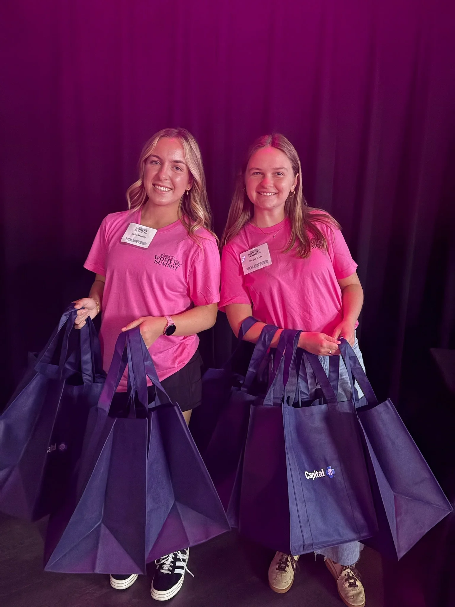 Two young women in pink shirts smiling, holding large black tote bags with the Capital logo, standing in front of a black curtain at an event.