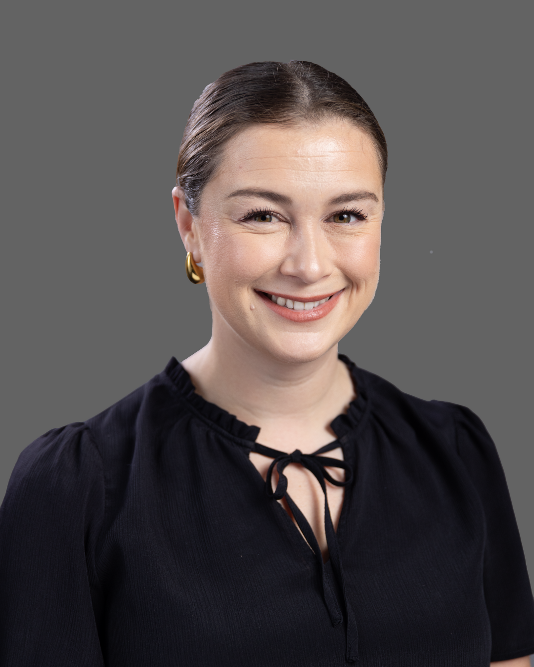 Smiling woman with brown hair pulled back, wearing gold hoop earrings and a black top with a tied neckline, against a gray background.