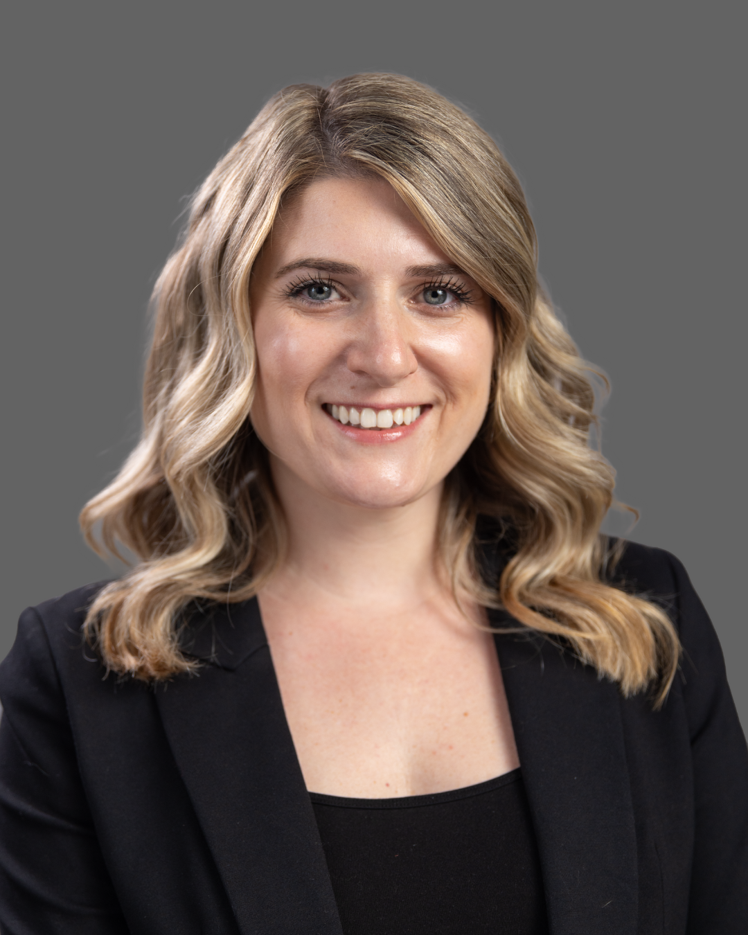 Portrait of a smiling woman with wavy blonde hair, wearing a black blazer, against a gray background.