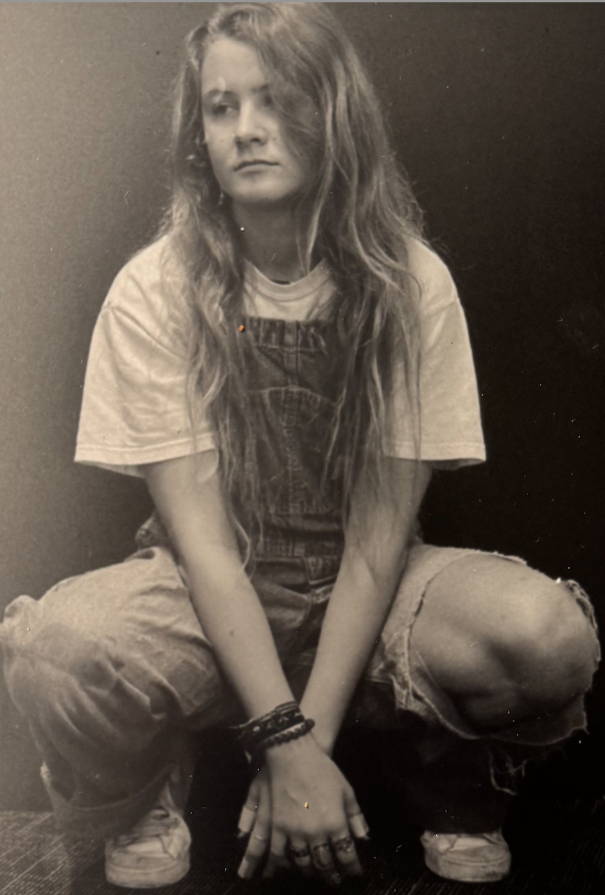 Black and white photo of a young woman with long, wavy hair, sitting cross-legged against a dark background, wearing a T-shirt, overalls, sneakers, and multiple bracelets.
