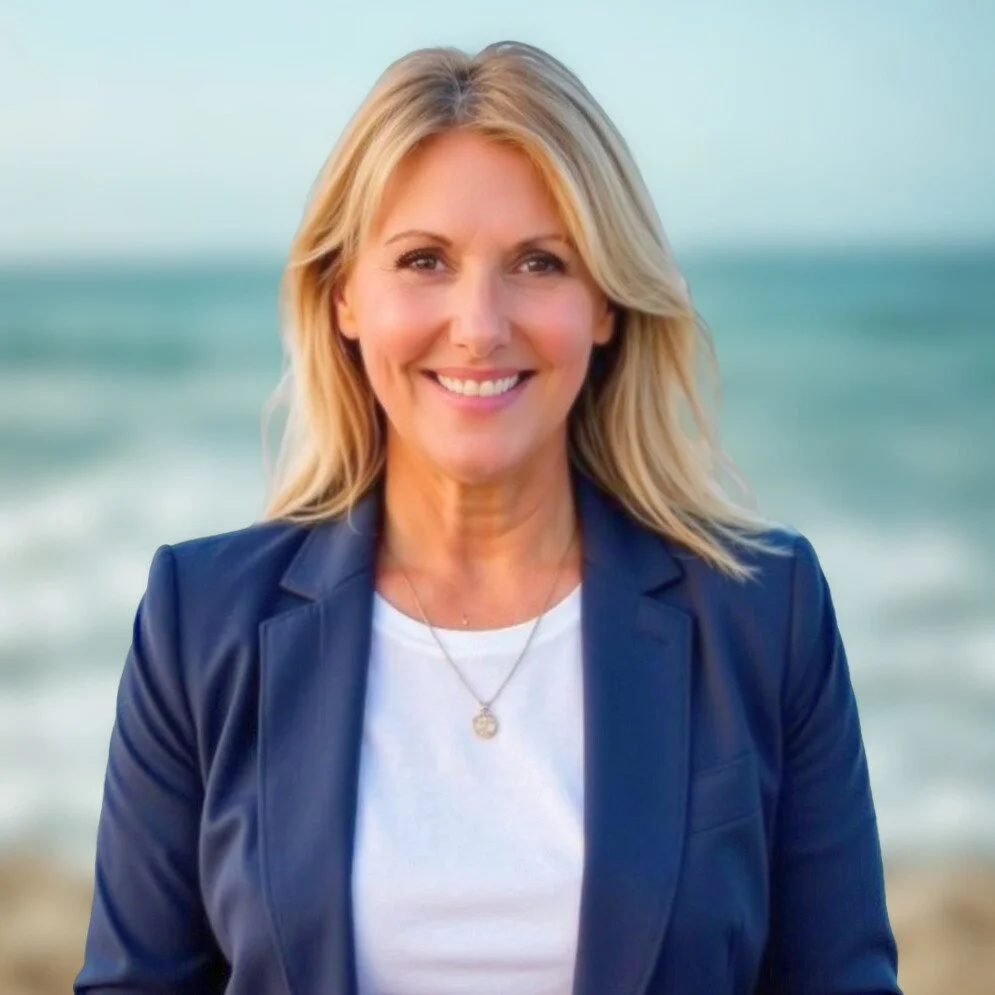 A smiling woman with blonde hair wearing a navy blazer and white top, standing on a beach with the ocean in the background.